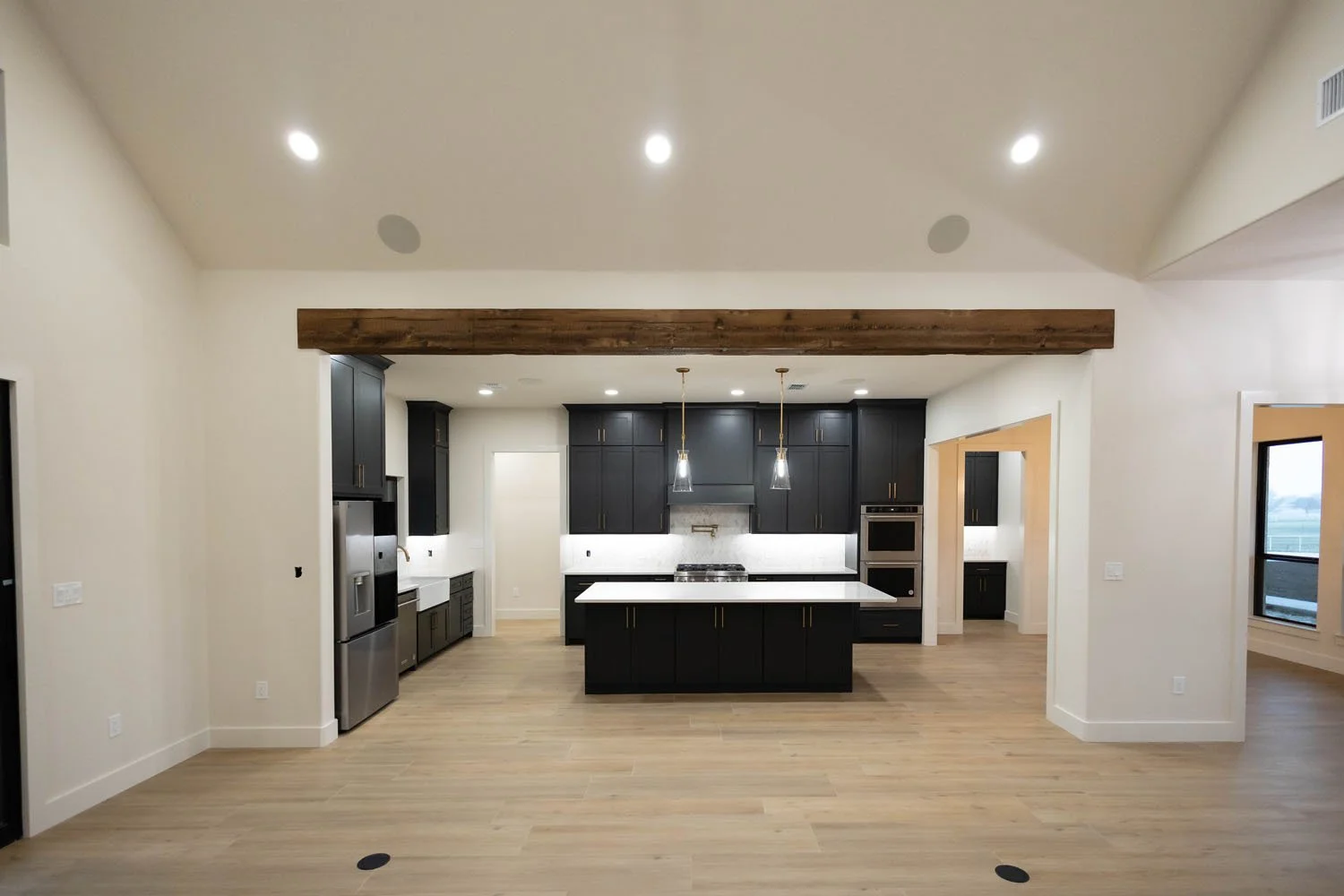 An open-concept living area featuring a large wooden ceiling beam, a central kitchen with black cabinetry and a white island, and light-colored wood plank flooring.