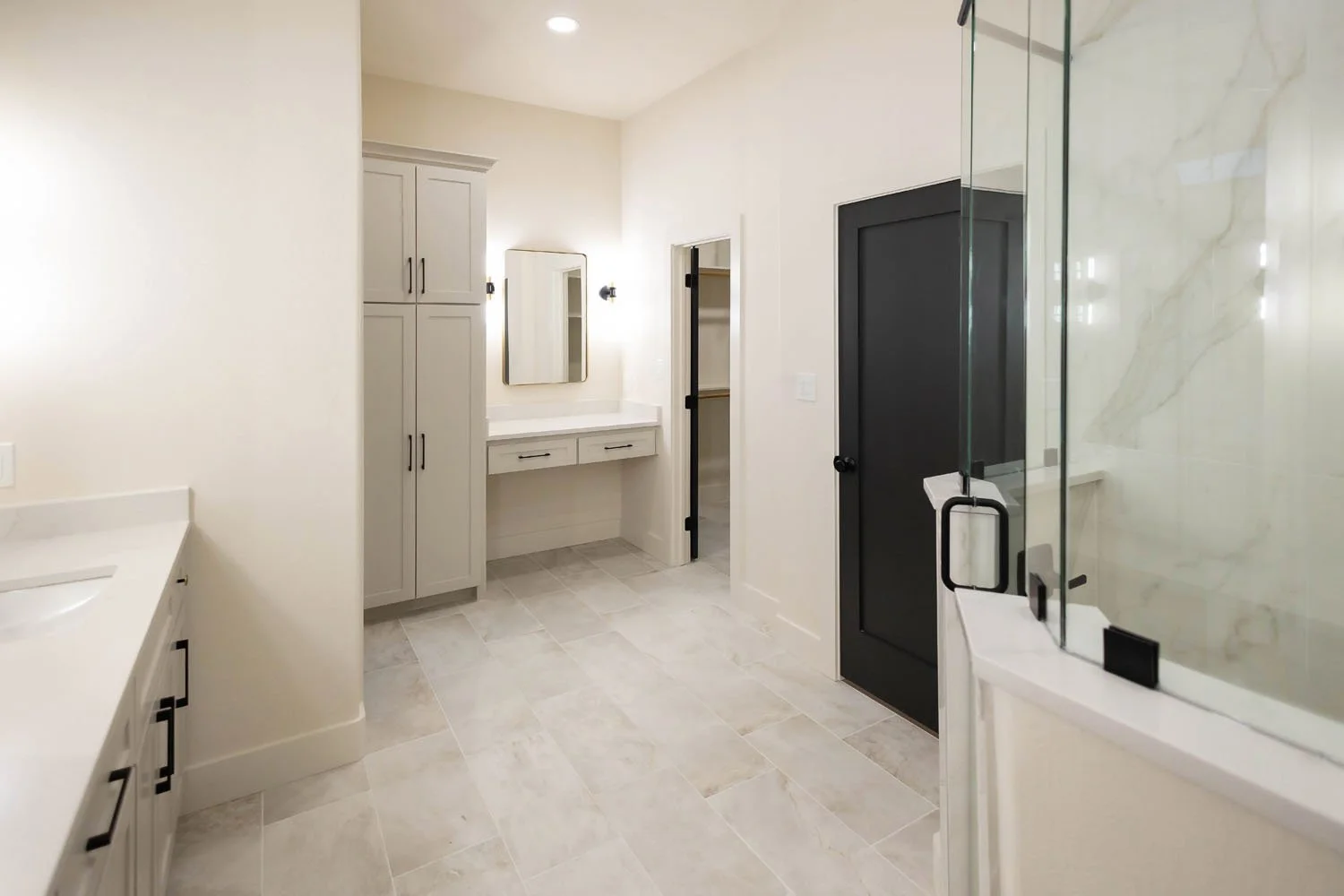 A bright bathroom featuring a white vanity, a tall linen cabinet, a small vanity desk with a mirror, and a glass-enclosed shower with marble-look tile.
