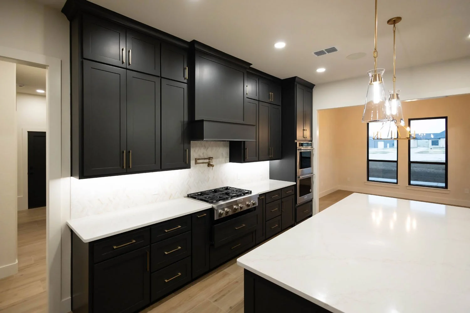 A kitchen interior with black cabinets, gold pendant lighting, a large white kitchen island, and a stainless steel range and double oven.