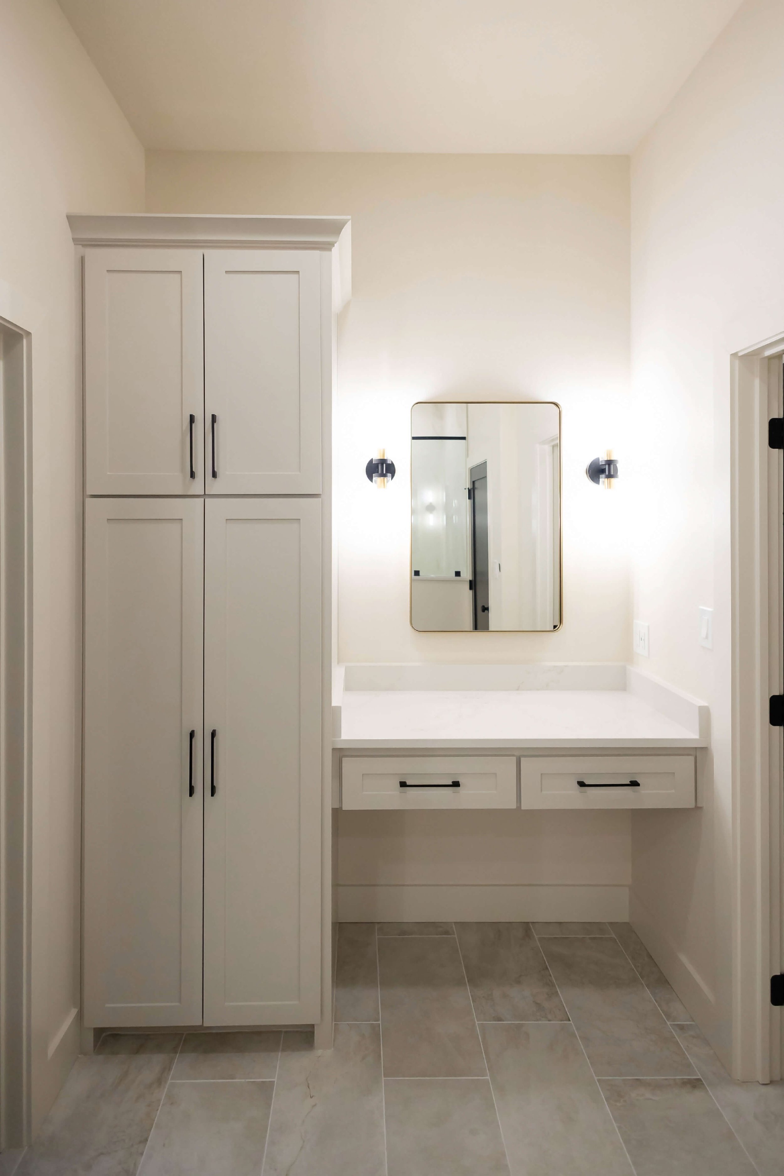 A clean, minimalist bathroom vanity featuring a light-toned tall cabinet, white countertop, rectangular mirror, and warm wall sconce lighting.