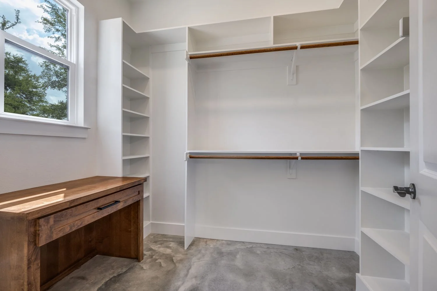 A modern, clean closet space featuring white shelving, wooden clothing rods, a built-in wooden desk, and a polished concrete floor.