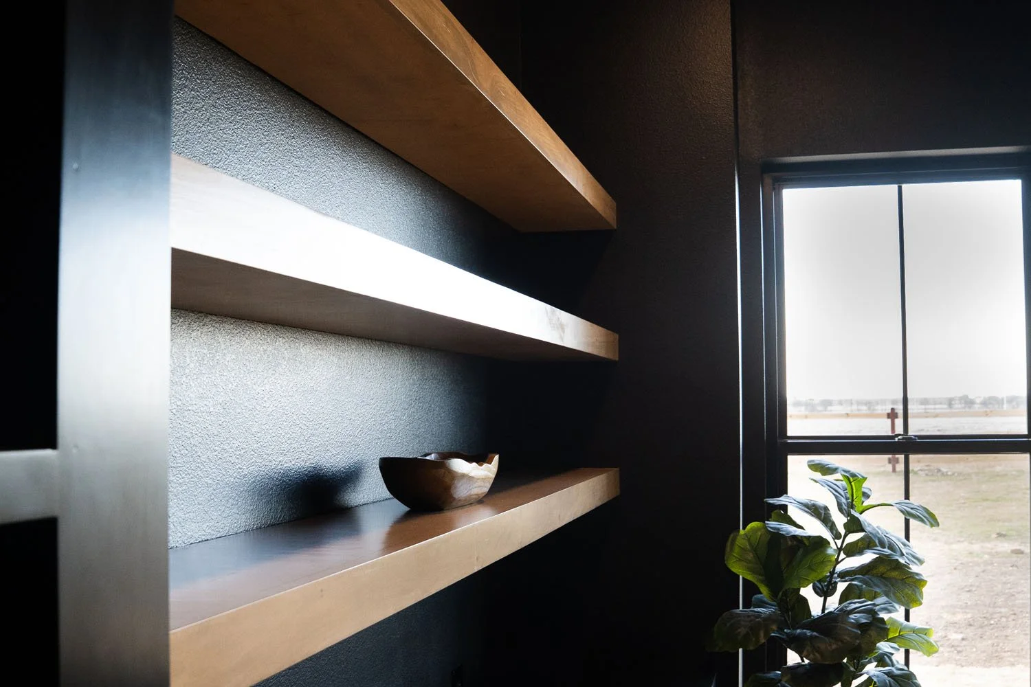 A close-up of dark charcoal walls in a modern office space, featuring three sleek floating light-wood shelves holding a wooden bowl, adjacent to a large window and a green potted plant.