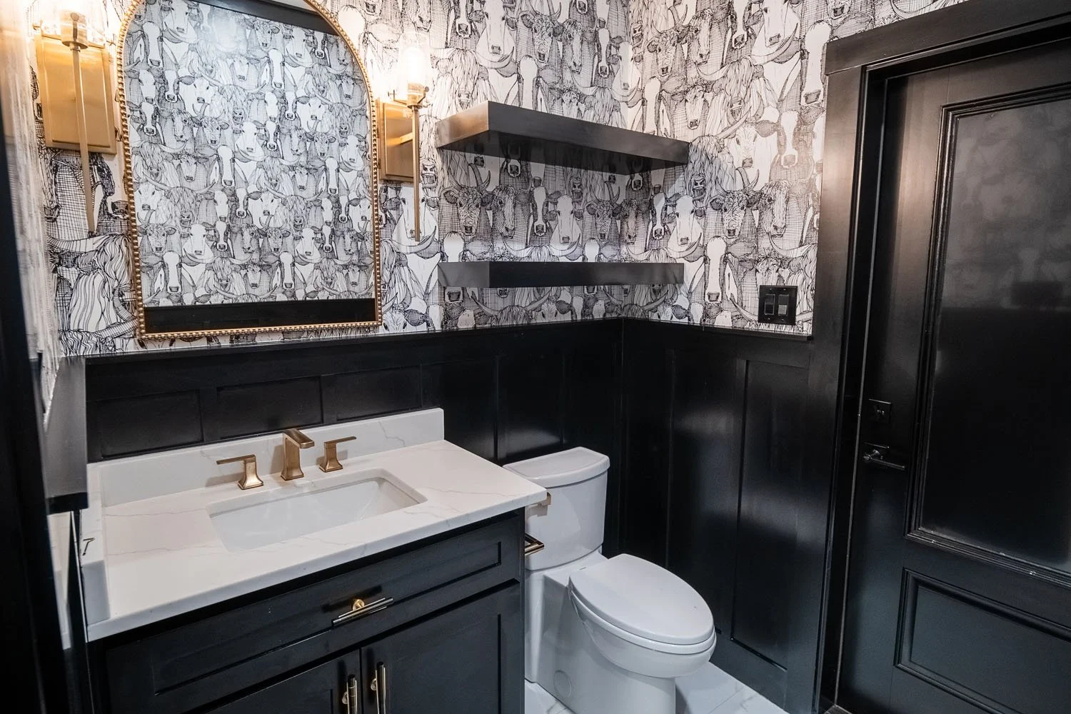 A stylish powder room featuring bold black and white cow-patterned wallpaper, a black vanity with a white quartz countertop, gold fixtures, and a black wainscoted wall.