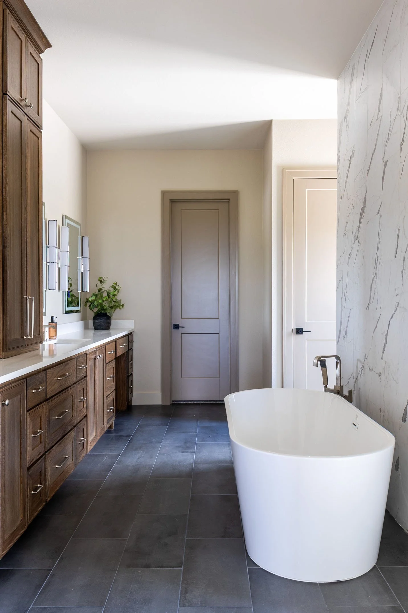 Modern bathroom with a white freestanding bathtub, dark tiled floor, wooden cabinets, a potted plant, and a windowed wall with marble accents.