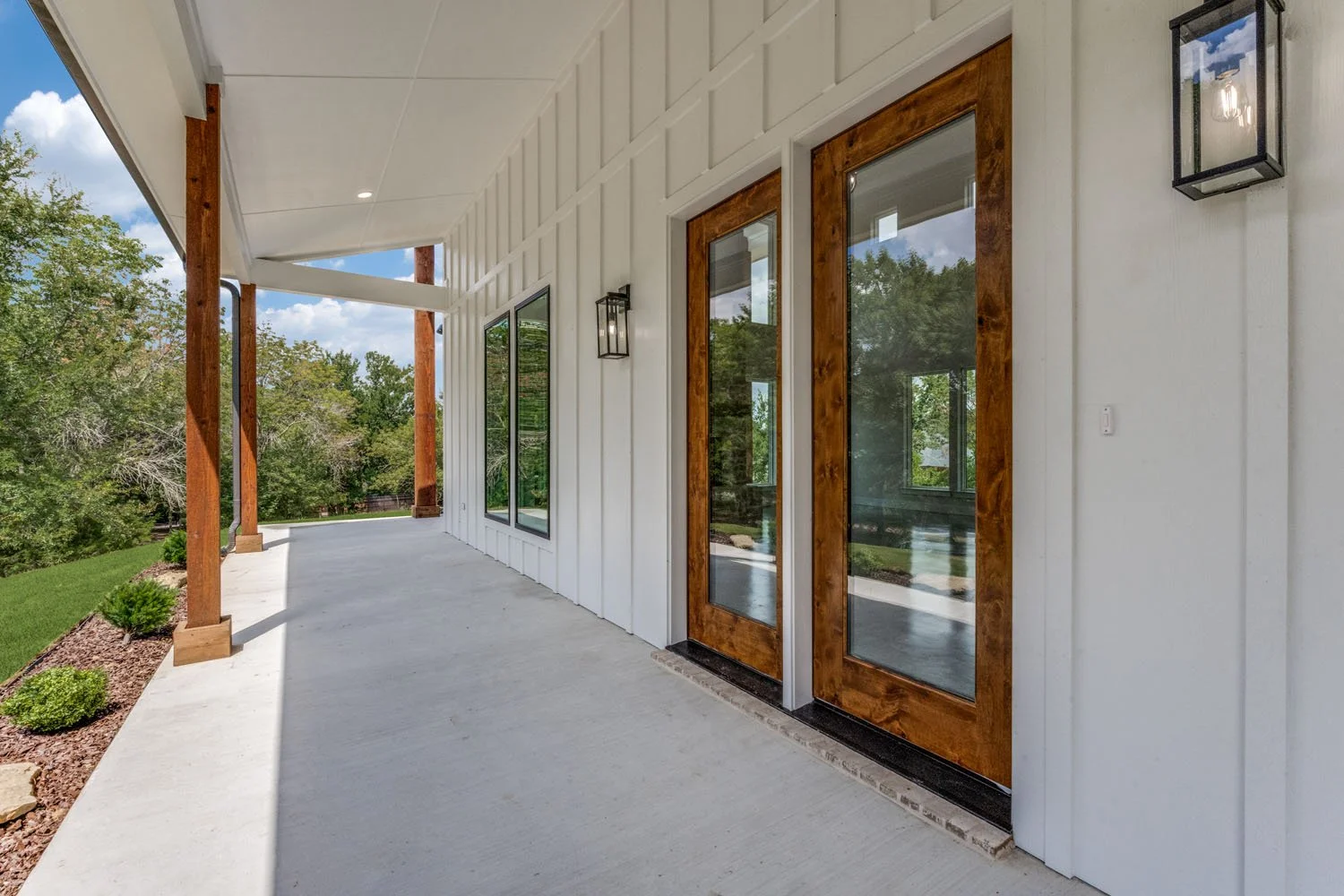 A close-up view of a covered porch featuring stained wood support columns, matching wood-framed glass doors, and black outdoor sconce lighting.