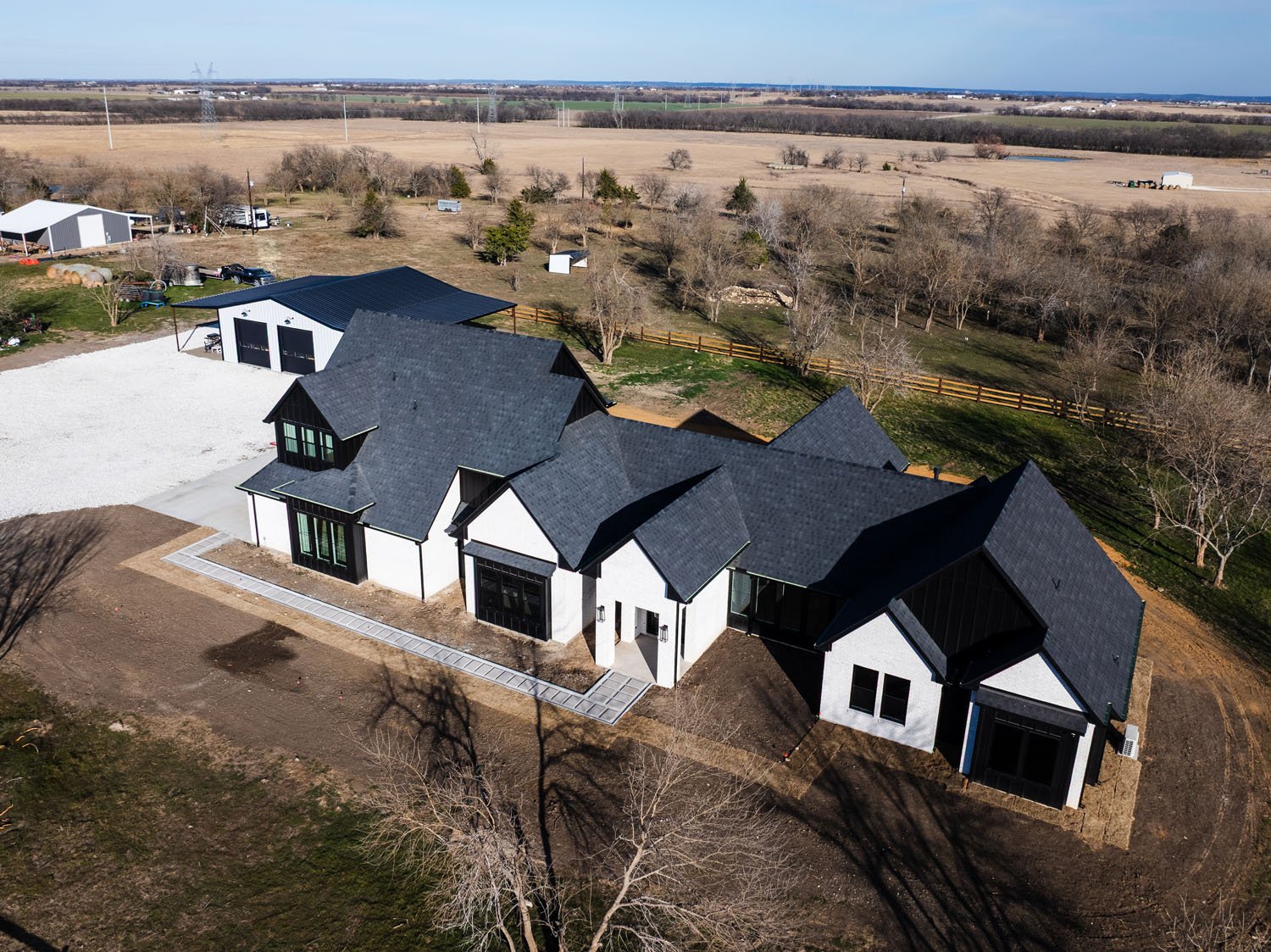An elevated drone shot of a custom farmhouse-style home set in a rural landscape, featuring a white brick and black vertical siding exterior, a multi-gabled roofline, and a detached metal workshop.