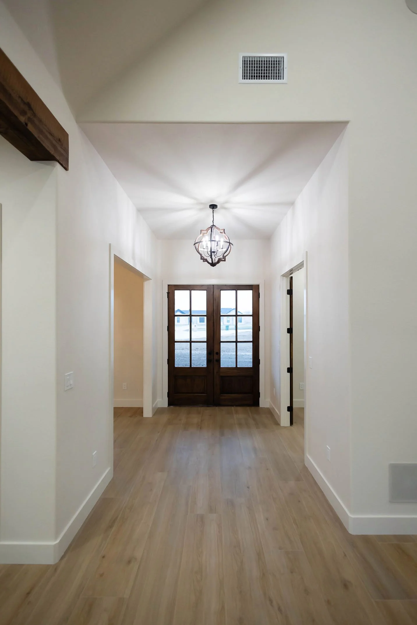 A brightly lit interior entryway featuring light wood floors, high ceilings with a decorative wooden beam, a geometric chandelier, and dark wood double front doors.