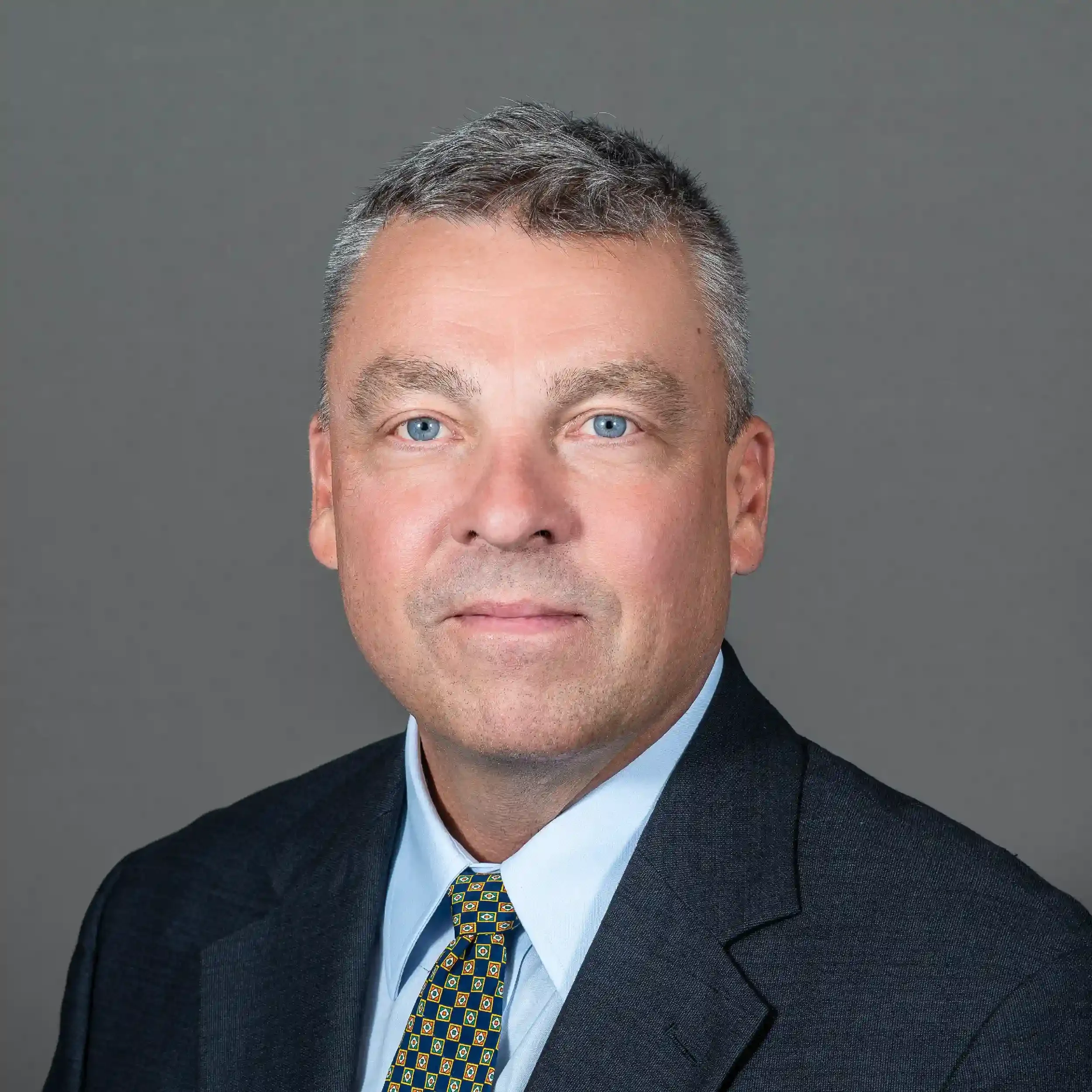 Close-up of a man with short graying hair, blue eyes, wearing a dark suit, light blue dress shirt, and a colorful patterned tie, against a plain gray background.