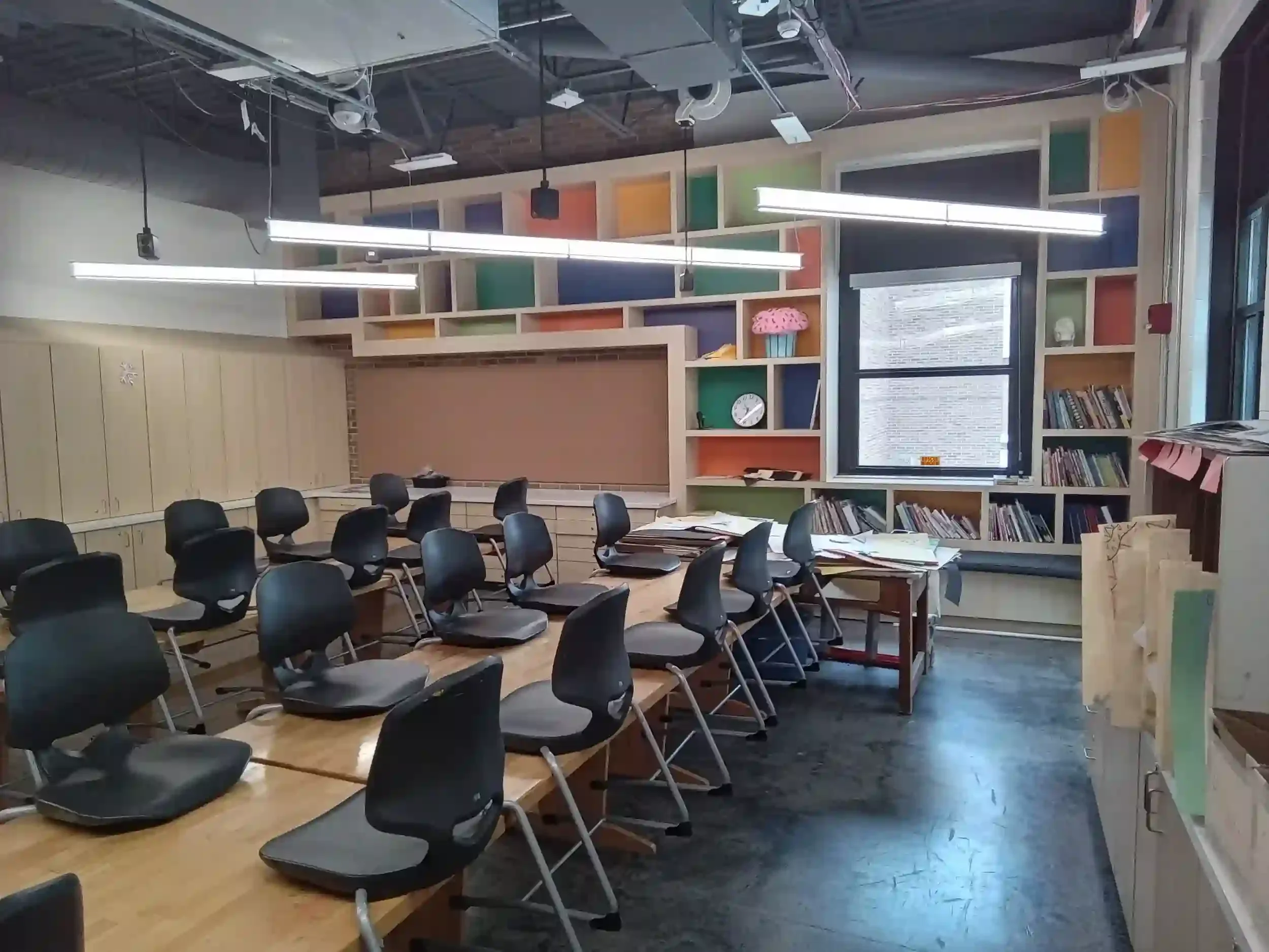 A classroom or meeting room with black chairs around rectangular wooden tables, colorful wall shelving, and a large window. The room has ceiling lights and visible air ducts.