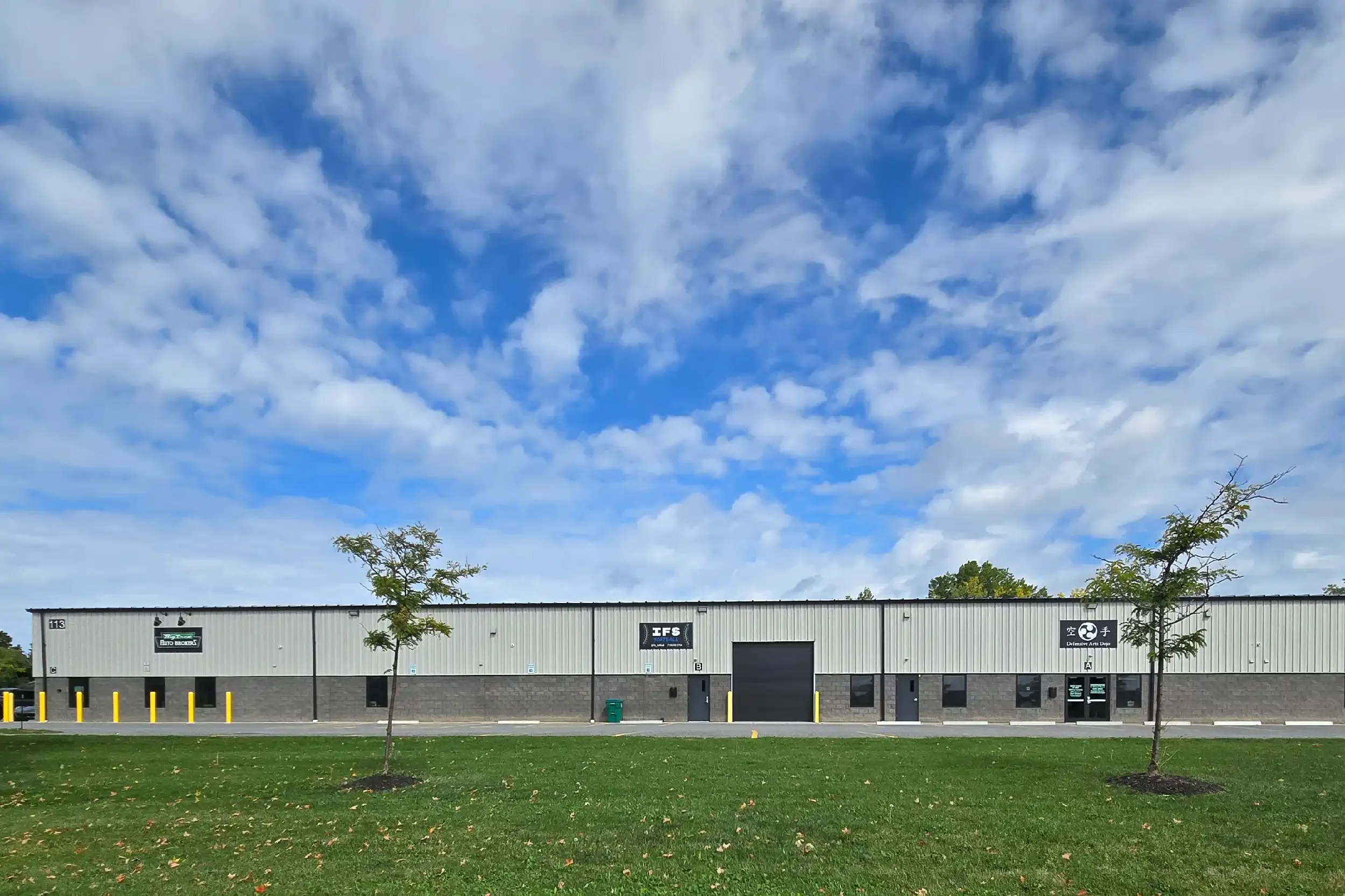 A large commercial building with several logos on its facade, surrounded by a green lawn with three small trees and a cloudy sky overhead.