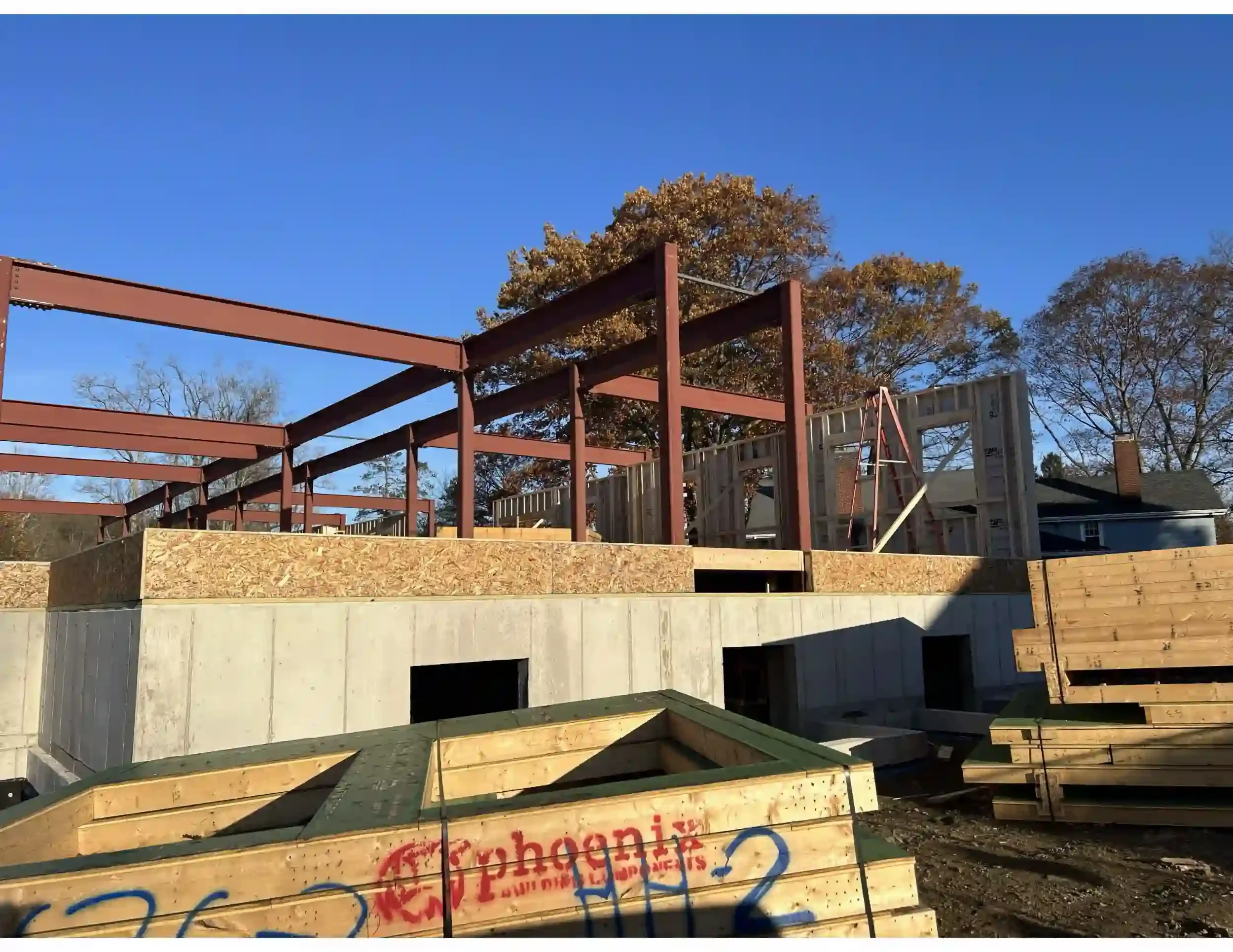 Construction site with framing structure and plywood walls, with blue sky and trees in the background.