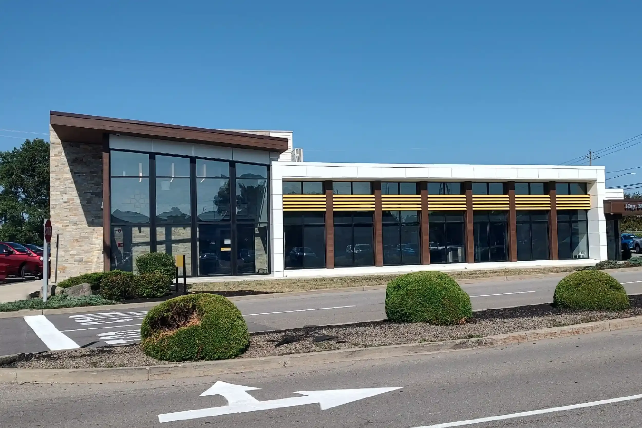 Modern commercial building with large glass windows and a combination of stone and white exterior design, located alongside a parking lot with a painted arrow on the road indicating the direction of traffic.