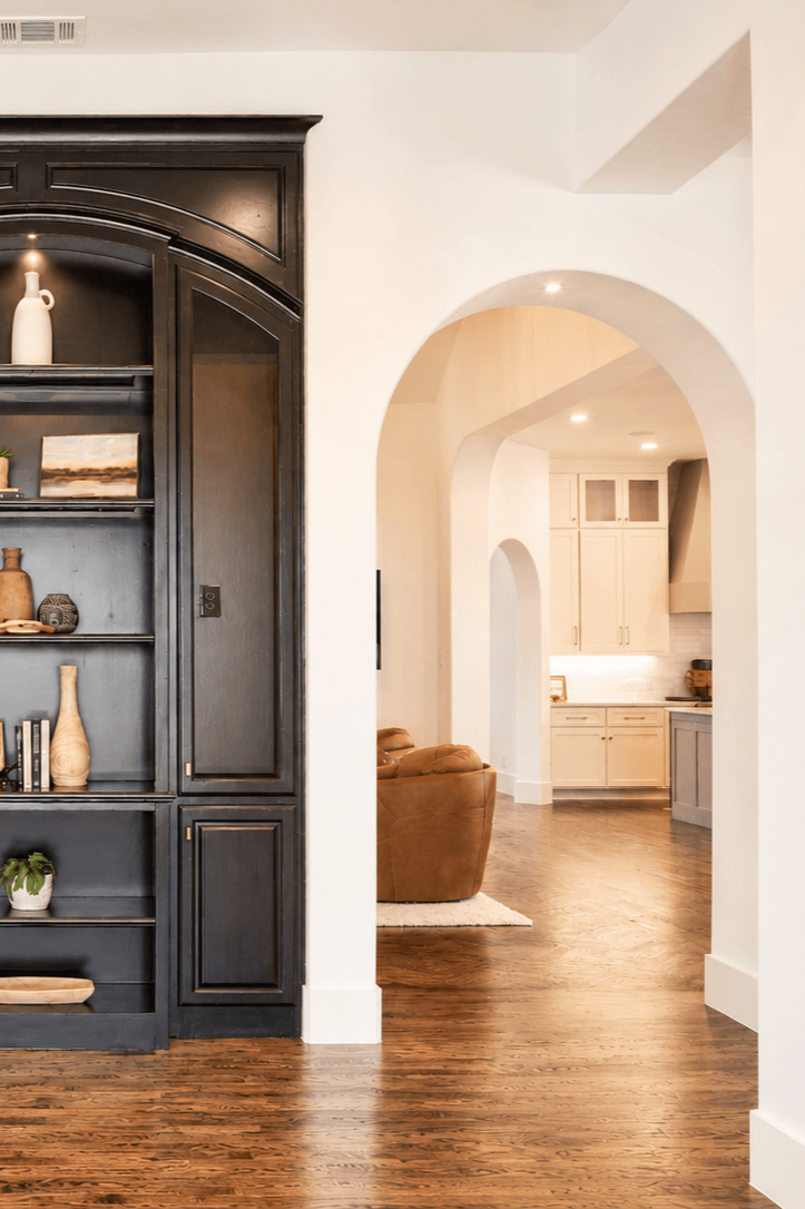 Living room with arched doorways leading to a kitchen, featuring a dark blue bookshelf, wooden flooring, a brown leather sofa, and white kitchen cabinets.