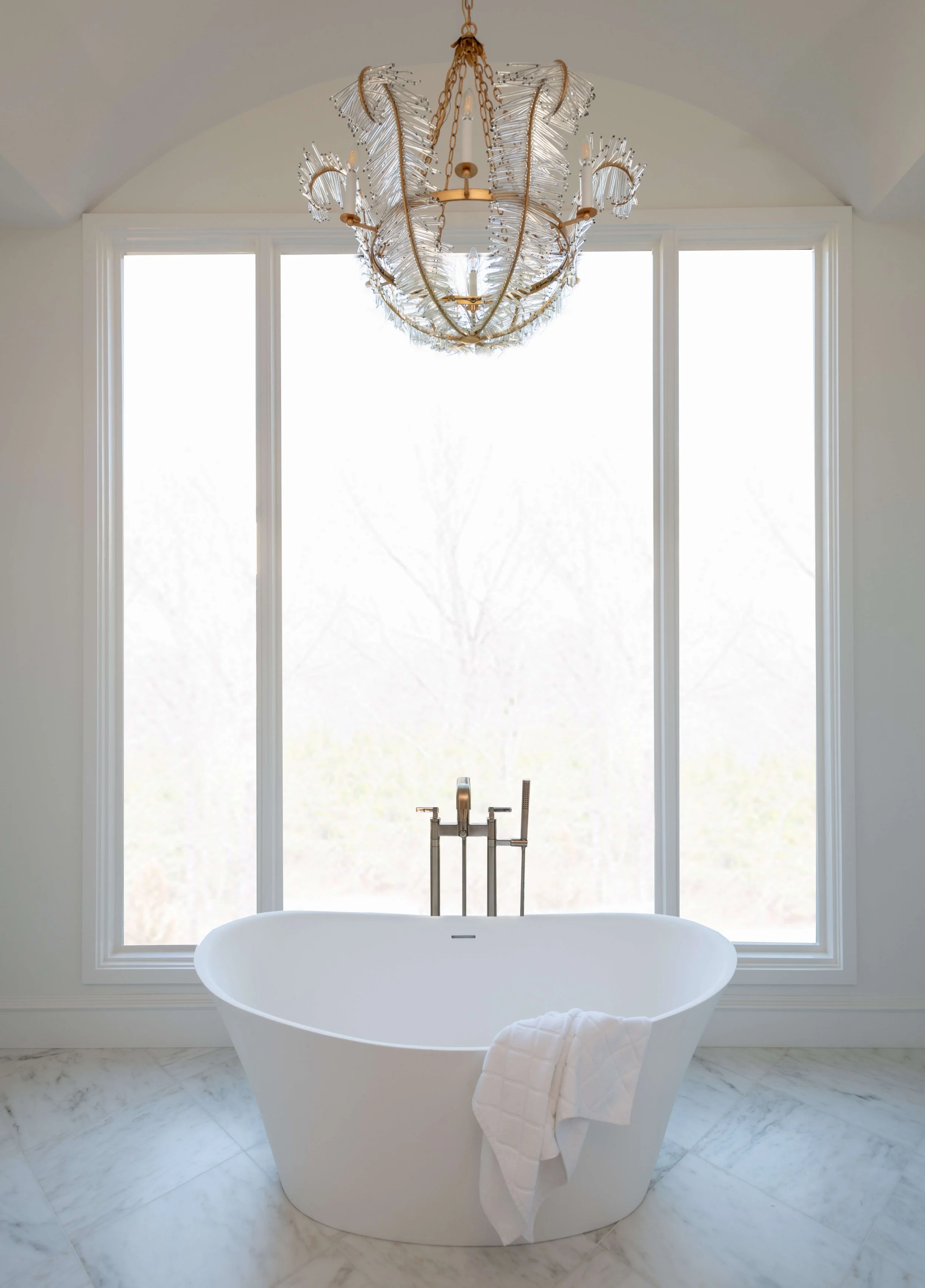 A modern bathroom with a white freestanding bathtub, positioned in front of large windows with frosted glass, a rose gold chandelier with glass details hanging from the ceiling, and a white towel draped over the edge of the tub, all with a marble floor.