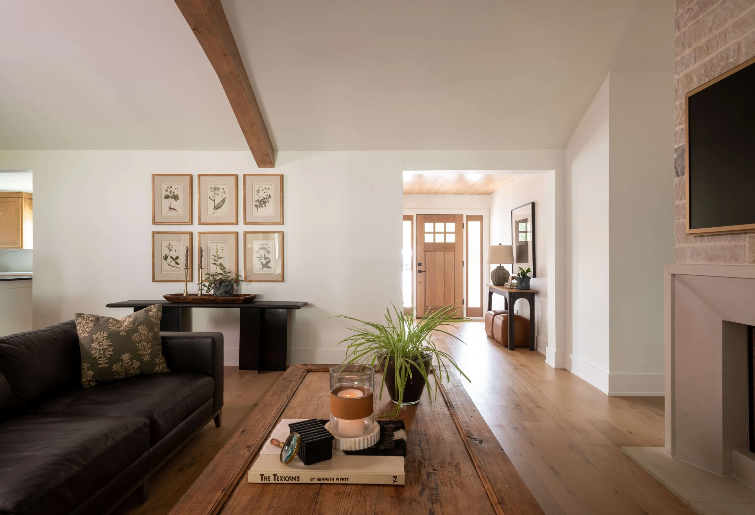A cozy living room with a black leather sofa, a wooden coffee table with candles and decorative items, and framed botanical artwork on a white wall. The room features hardwood floors, a front door with a window, and natural light.