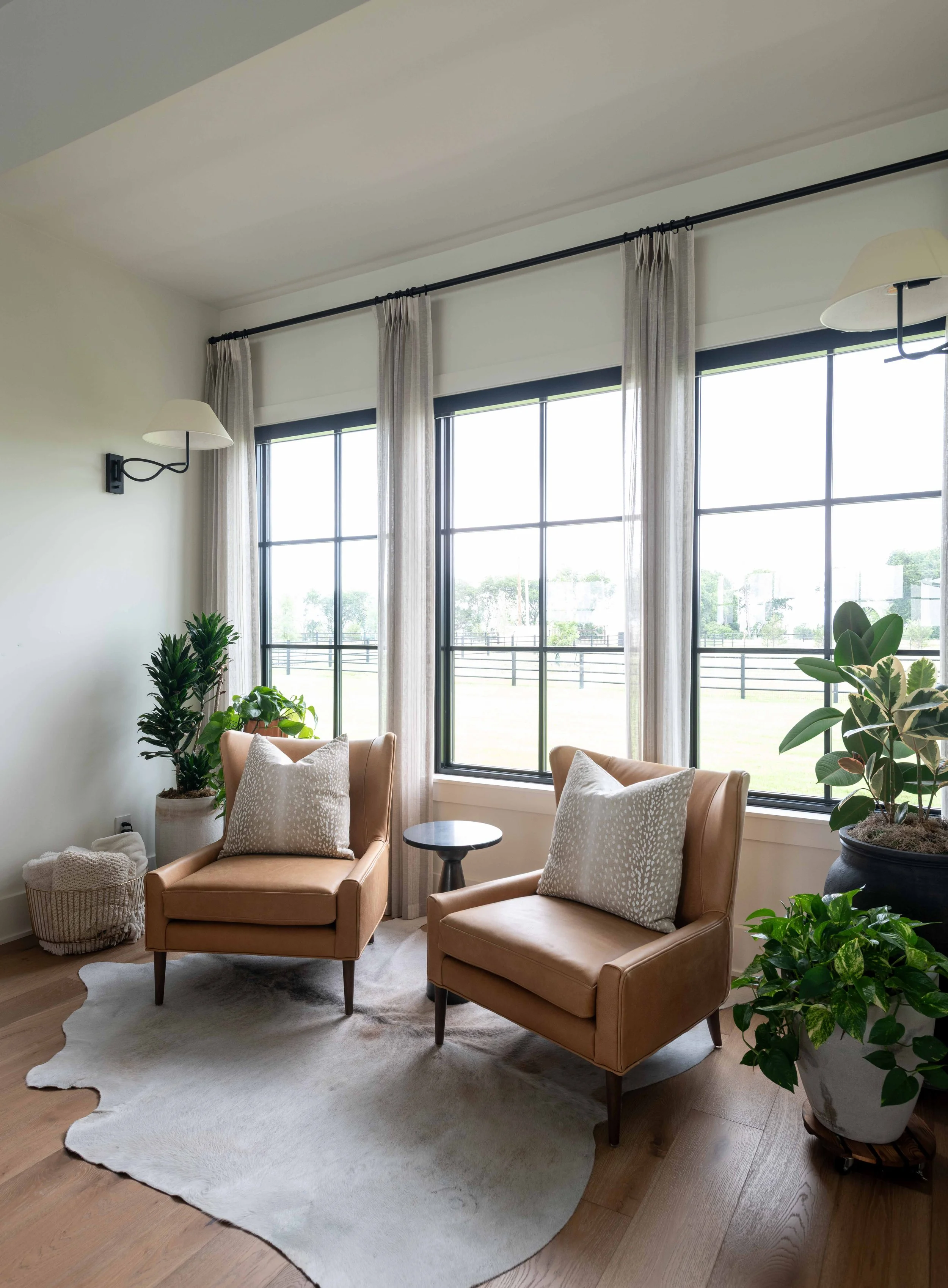 Living room with large windows, beige chairs with patterned pillows, potted plants, and a white rug.