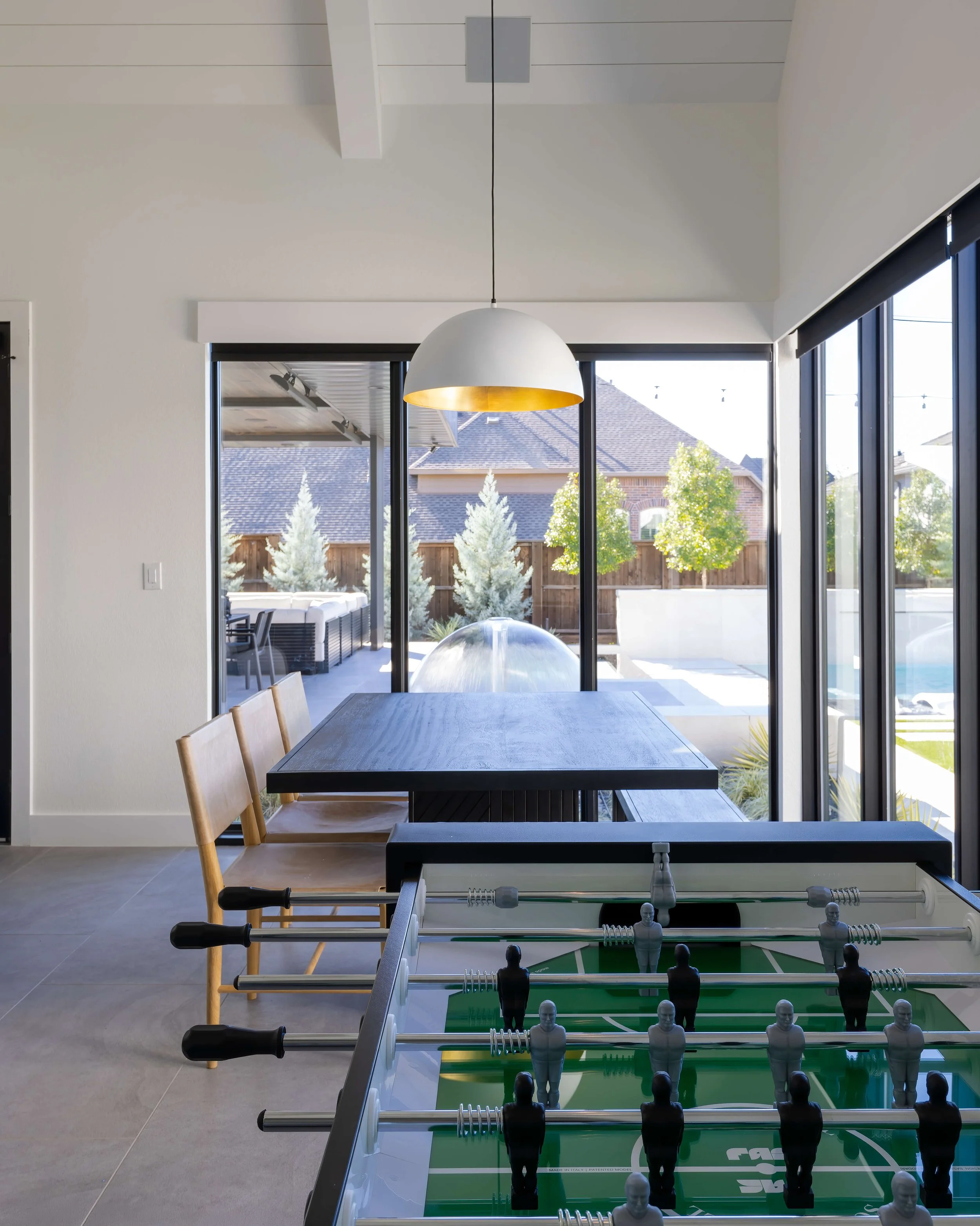 View of an indoor recreational room with a foosball table in the foreground, a table and chairs, and large sliding glass doors leading to an outdoor patio with trees and houses.