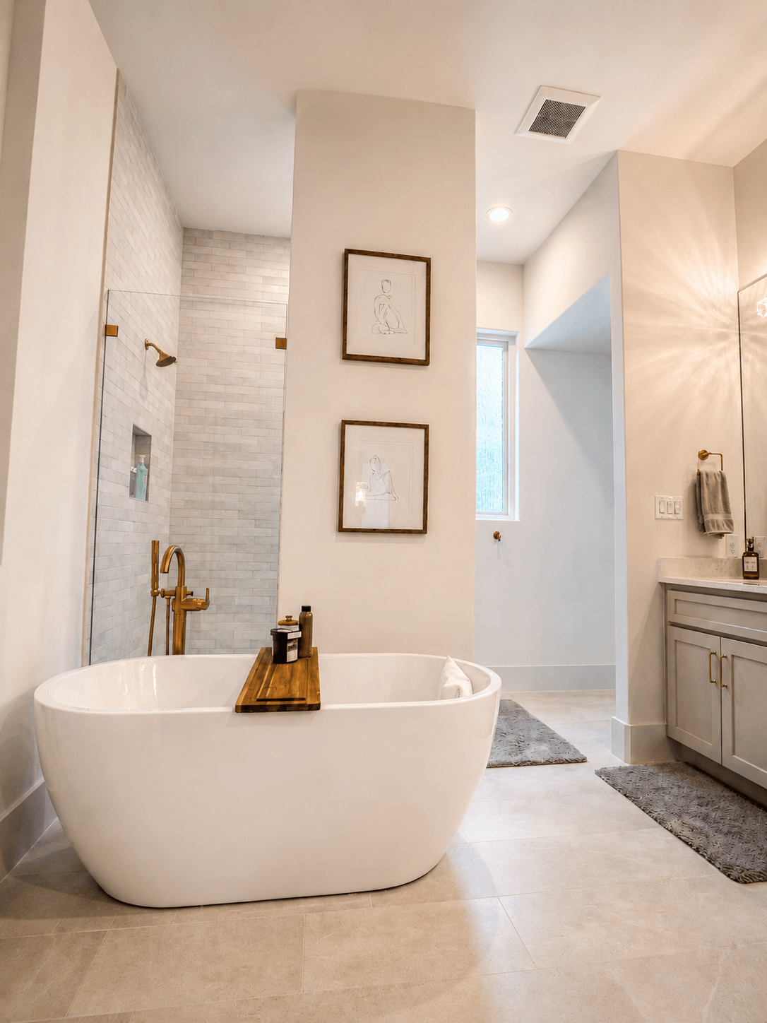 Modern bathroom with a freestanding bathtub, each with a wooden tray and seating, a walk-in shower with white brick tiles, framed minimalist artwork, a window, and a vanity with a mirror, towel, and gold fixtures.