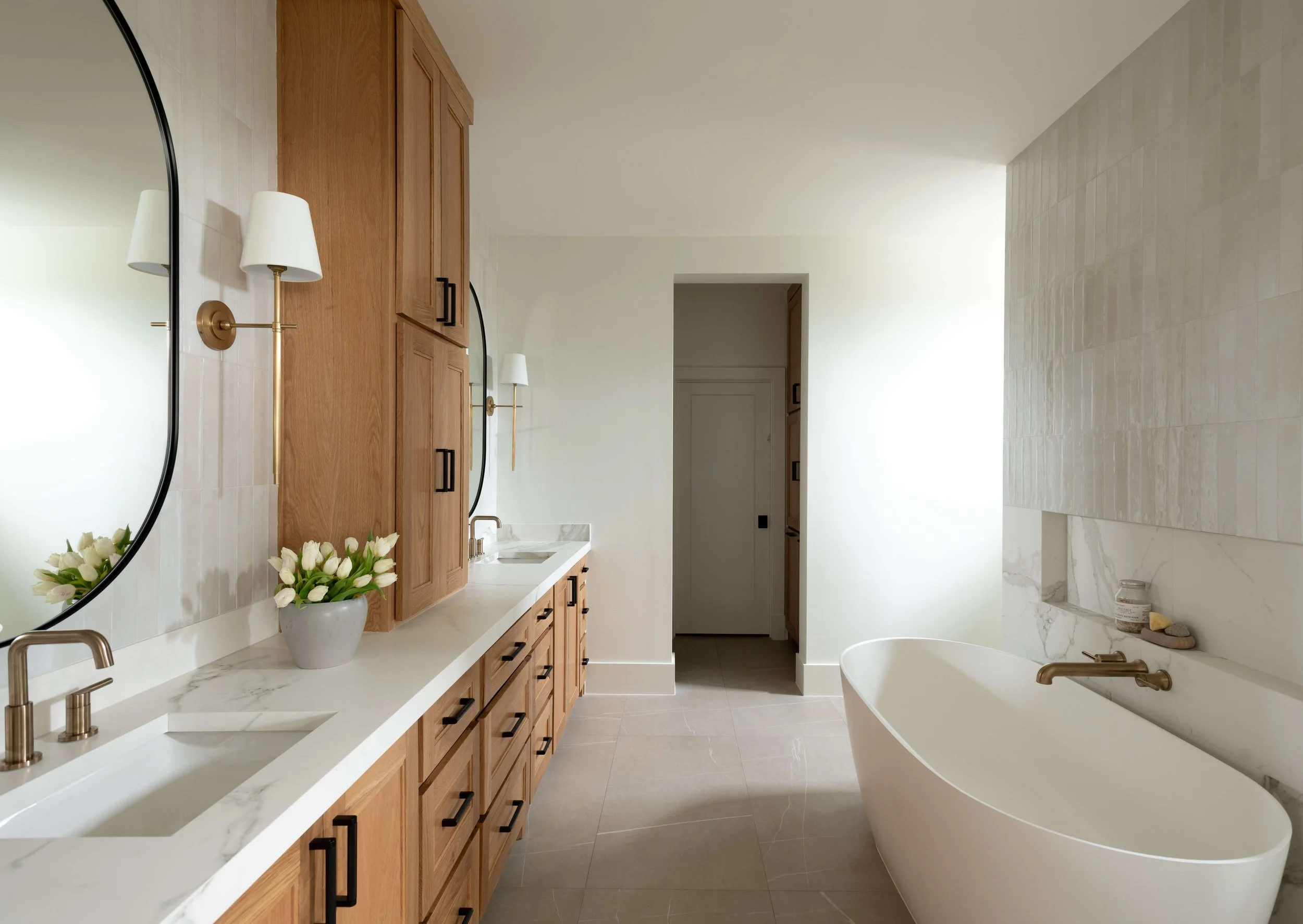 Modern bathroom with wooden cabinets, white marble countertop, oval mirror, brass wall sconces, vase with white tulips, white bathtub with brass faucet, tiled wall, and neutral-colored floor tiles.