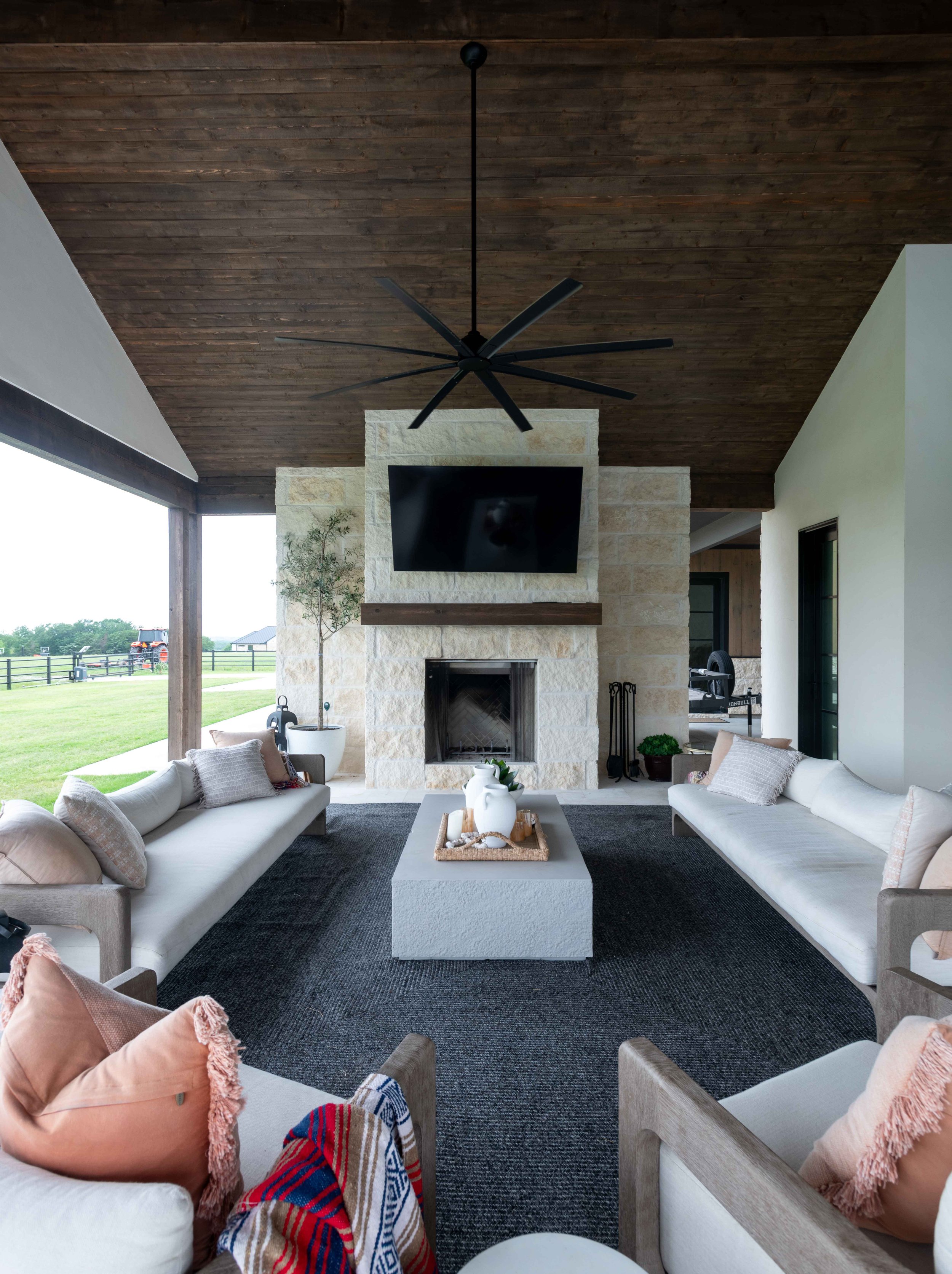 A covered outdoor living space with a wooden ceiling, stone fireplace, and a large flat-screen TV mounted above the fireplace. There are two white sofas with pillows, a black rug, and a rectangular coffee table with a white vase and decorative items.