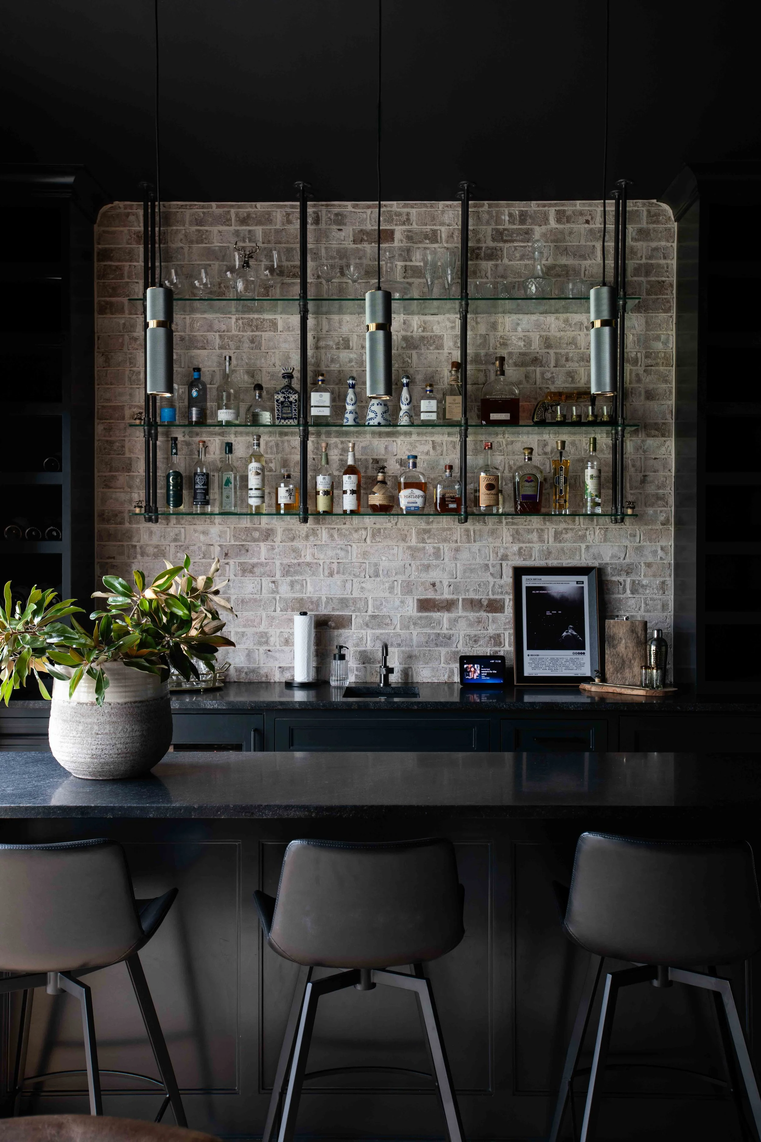 A modern bar with glass shelves holding liquor bottles and glassware, a brick wall background, a plant on the counter, and black chairs in front of a dark countertop.