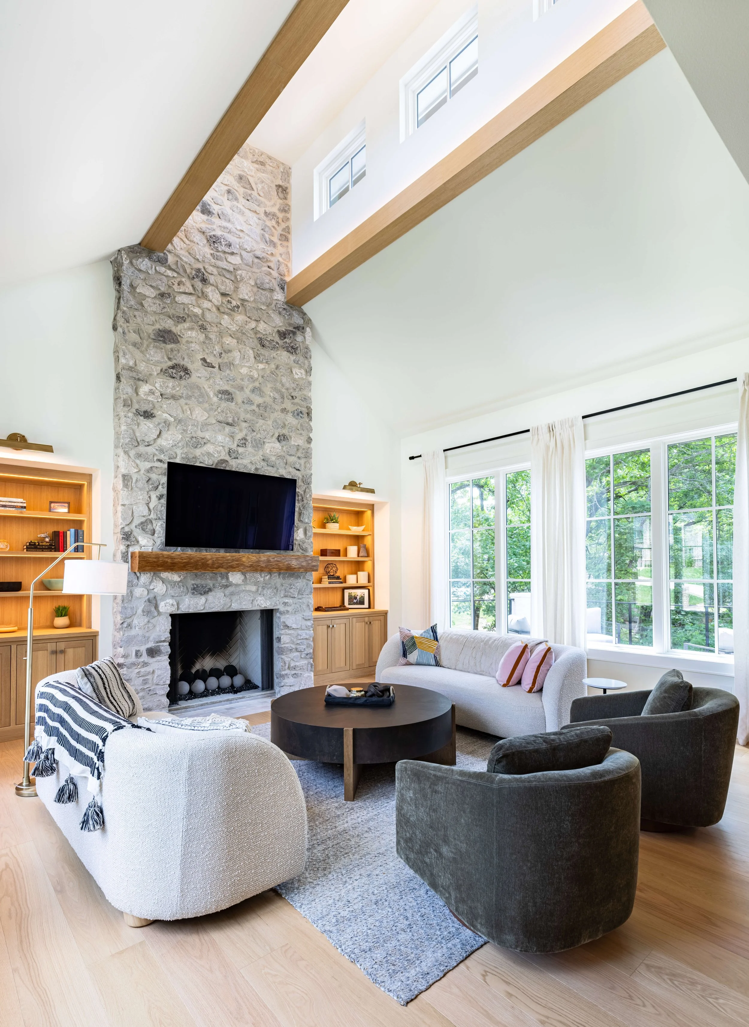 Living room with a stone fireplace, black TV, white sofa with pillows, a black round coffee table, and large windows with white curtains, bright natural light