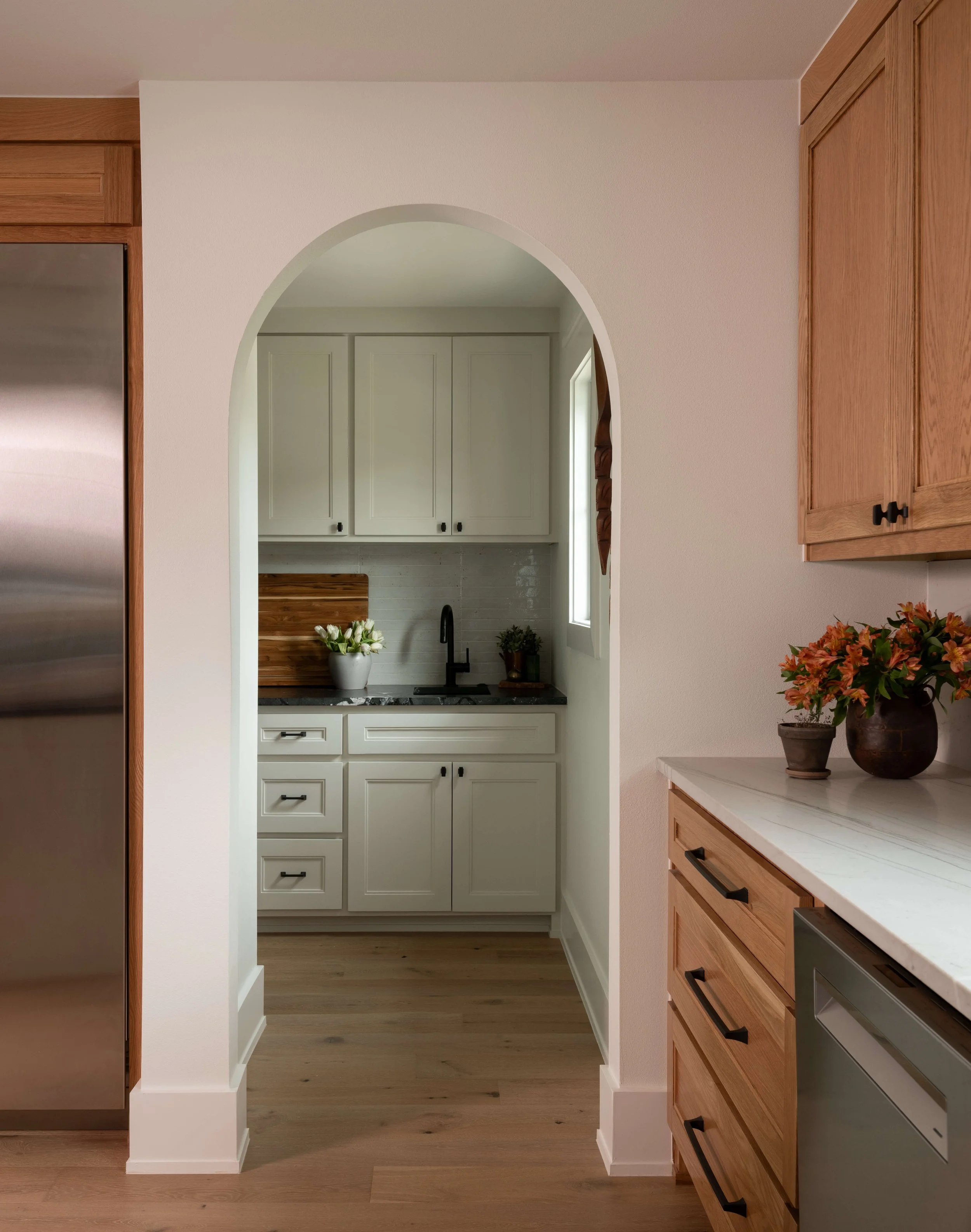 A view of a kitchen framed by a white archway, showing white cabinets, a black faucet, a wooden cutting board, a potted plant, and a bouquet of white flowers on the countertop, with wooden and stainless steel appliances and cabinets surrounding the space.
