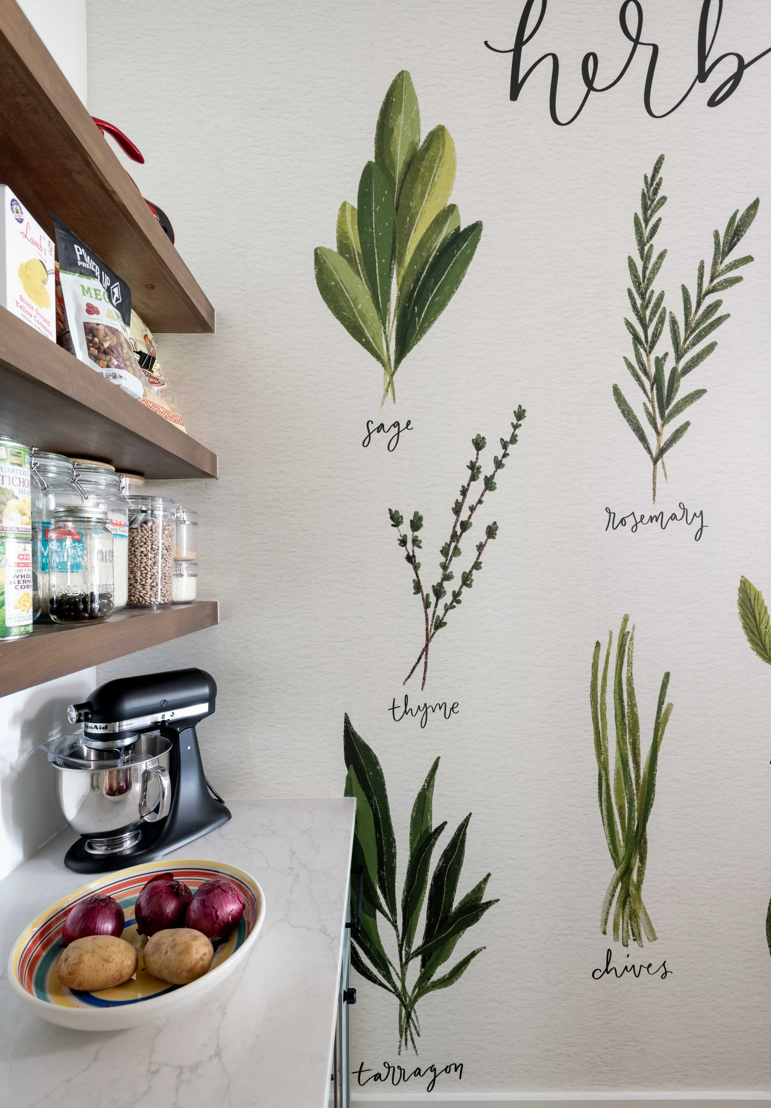 A kitchen countertop with a bowl of red onions and potatoes, a black stand mixer, and shelves with various food products. The wall behind has a painted herb garden with illustrations of sage, thyme, rosemary, chives, and tarragon labeled in cursive.