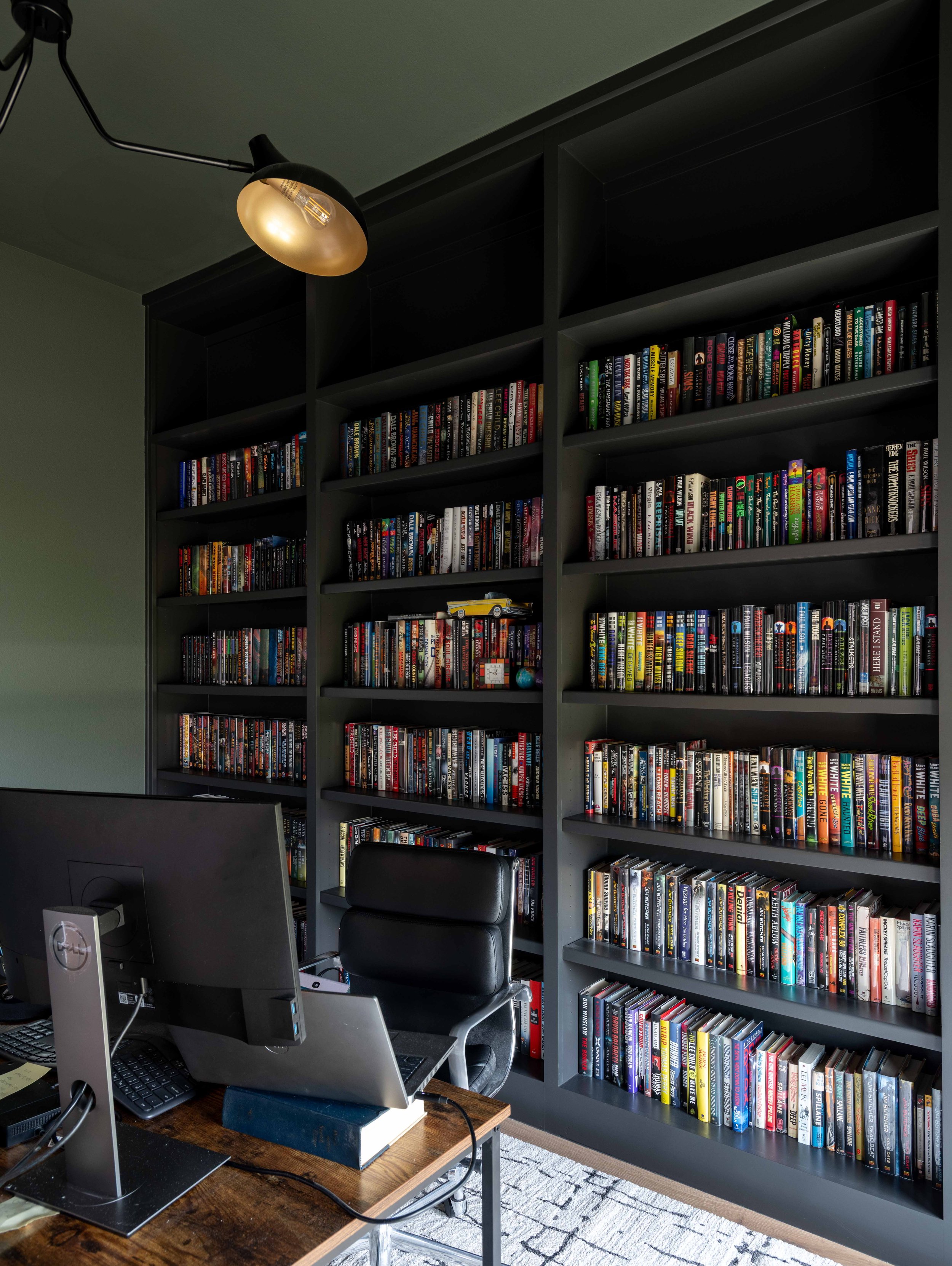 A home office with a large black bookshelf filled with numerous books, a computer monitor, keyboard, mouse, and a desk lamp. There is a black chair and a textured rug on the floor.