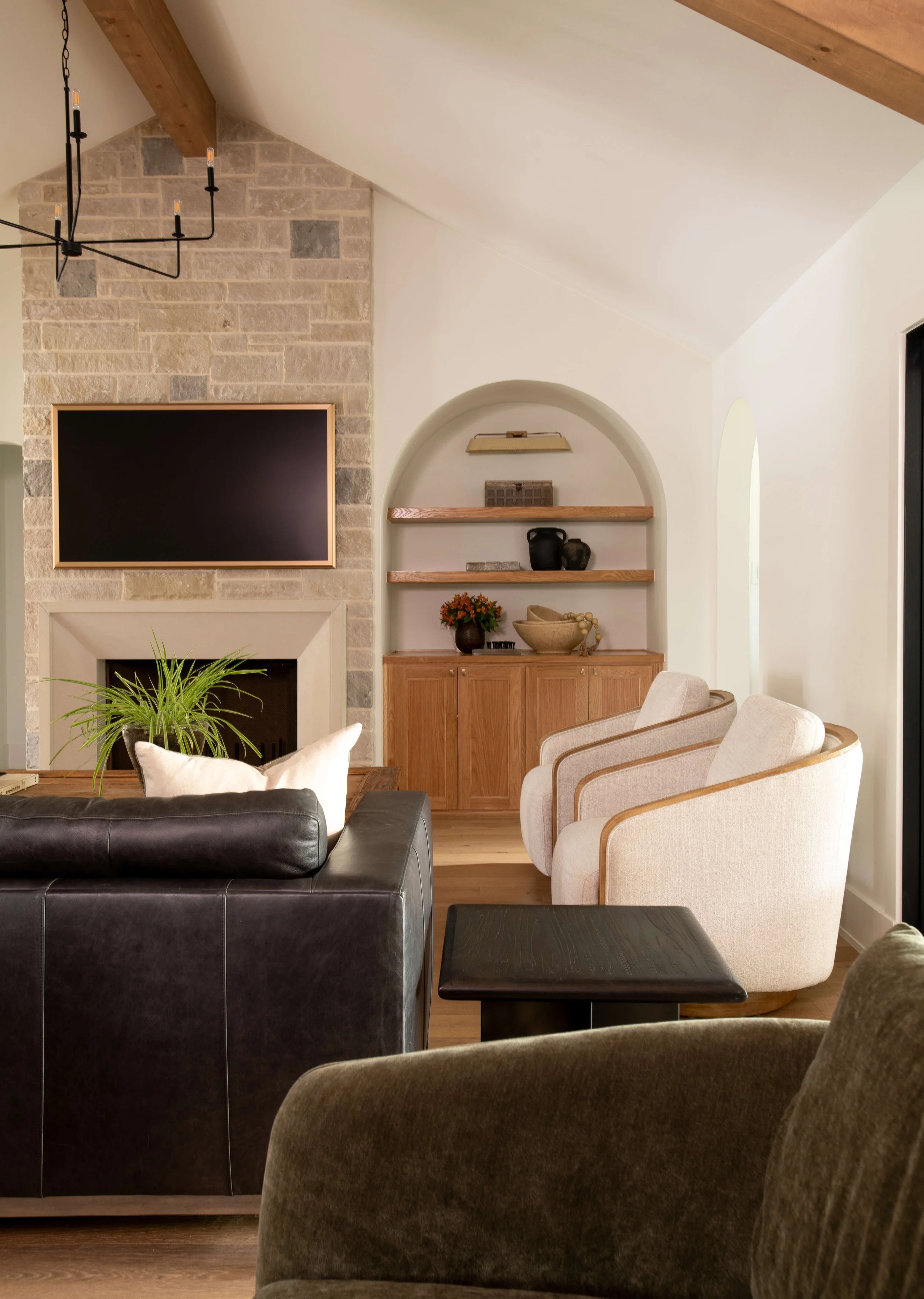 Living room with beige armchairs, a black leather couch, a fireplace with a stone surround, a wooden shelving unit with decorative items, and a black coffee table; part of a chandelier visible at the top.