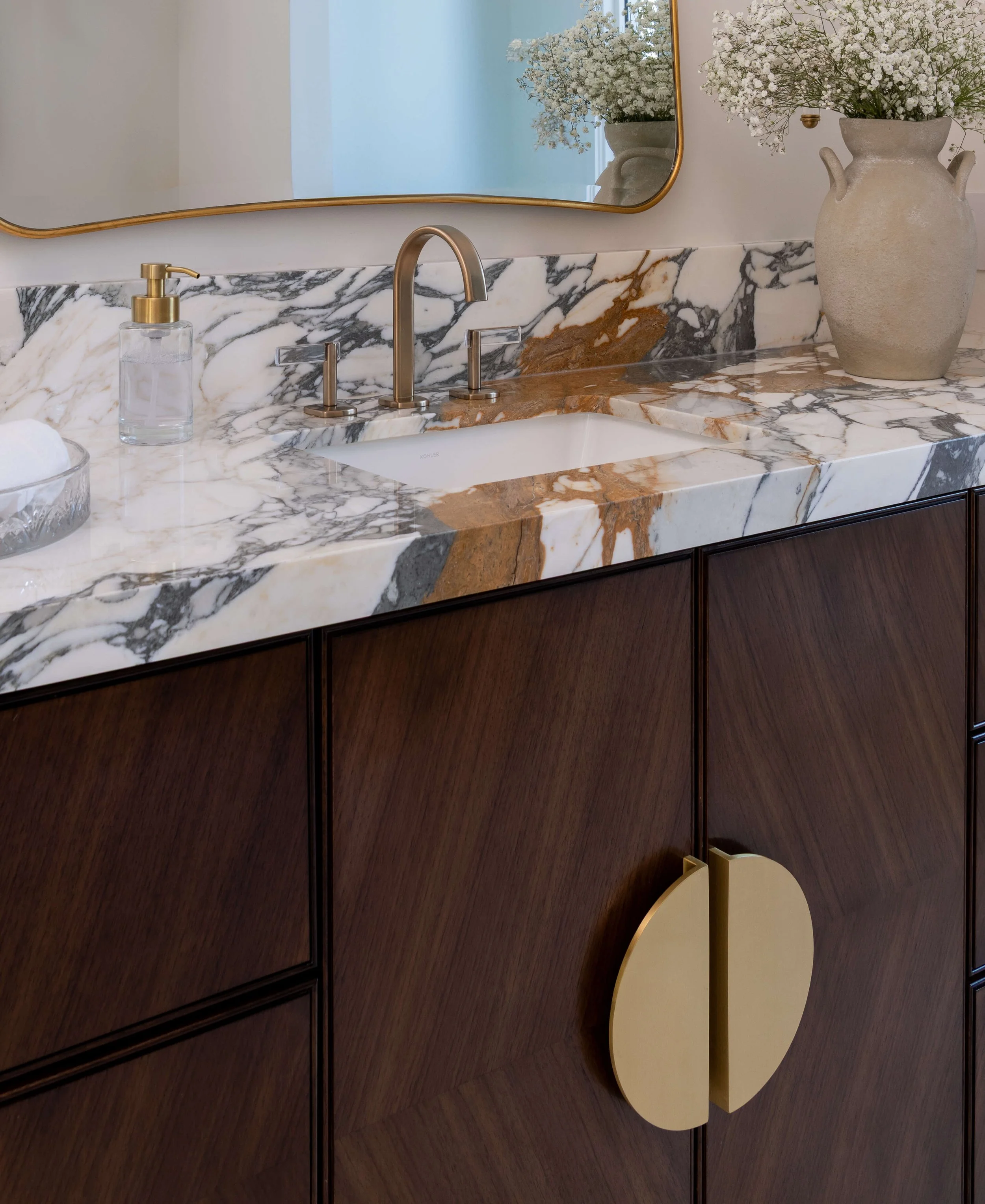 Bathroom vanity with marble countertop, gold faucet, and decorative vase with white flowers.