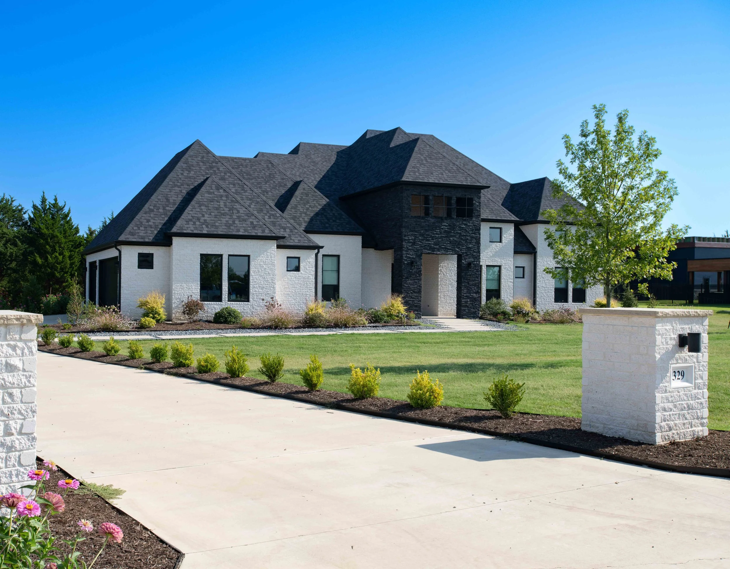 A modern house with a dark gray roof and white and black exterior walls, surrounded by a landscaped yard with green grass, bushes, and a sidewalk, under a clear blue sky.