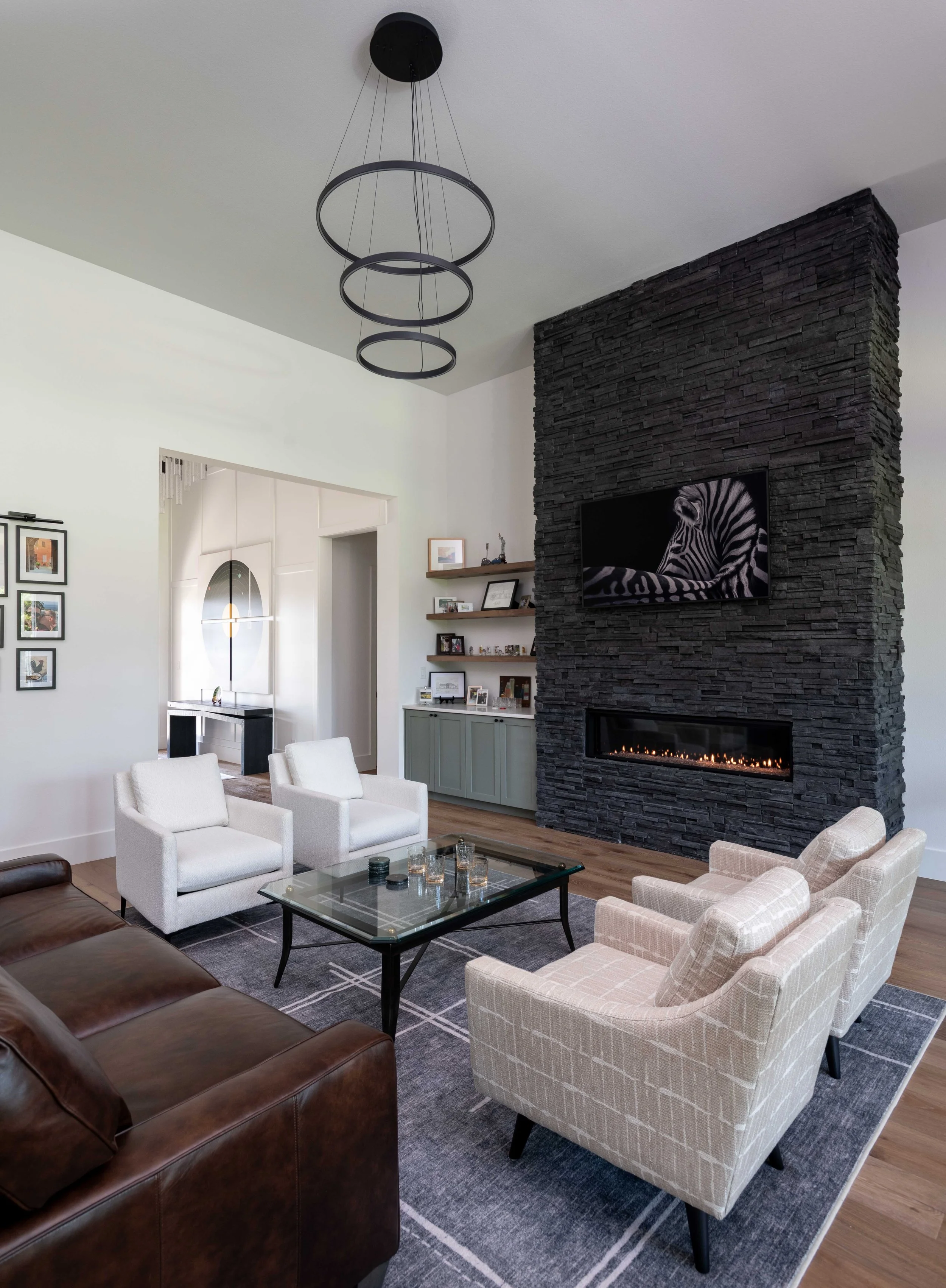 Living room with black stone fireplace, flat-screen TV with zebra image, white chairs, brown leather sofa, glass coffee table, patterned rug, and multiple wall-framed pictures.