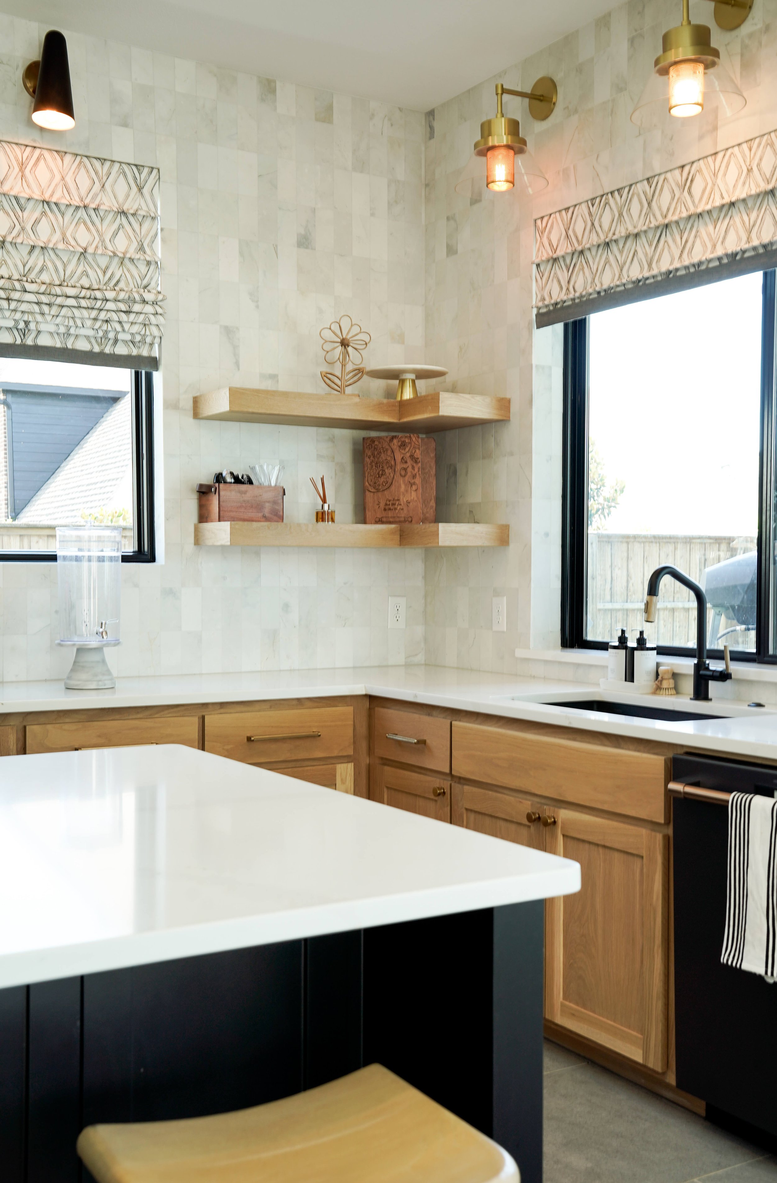 A modern kitchen with white countertops, wooden cabinets, and black accents. Two windows with geometric patterned curtains bring in natural light. Open wooden shelves display decorative items and kitchen accessories. Warm pendant lights hang from the ceiling, and a water dispenser is visible near the window.