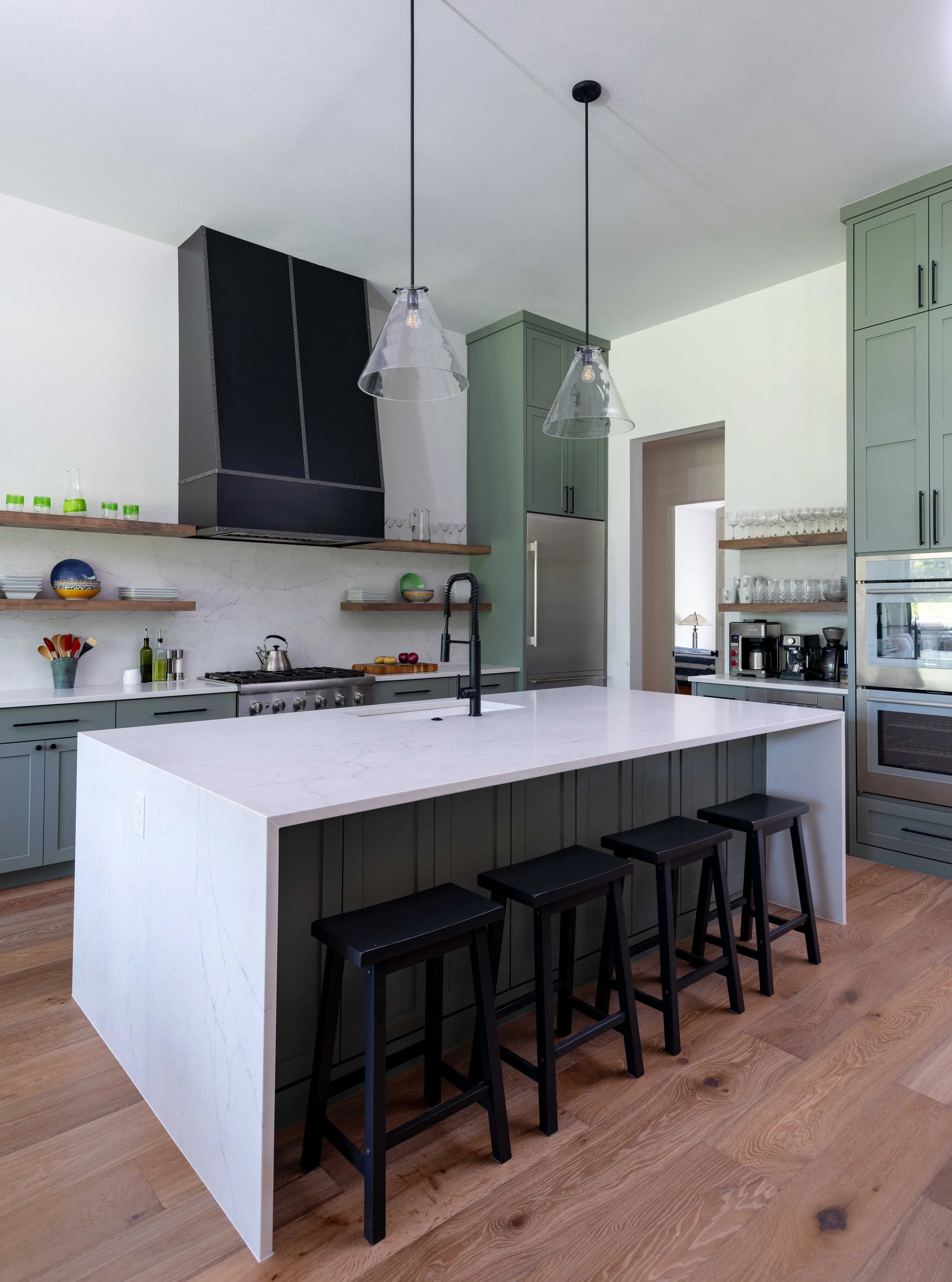 Modern kitchen with green cabinetry, a large white kitchen island, black stools, wooden shelves with dishes and glassware, and black pendant lights hanging from the ceiling.