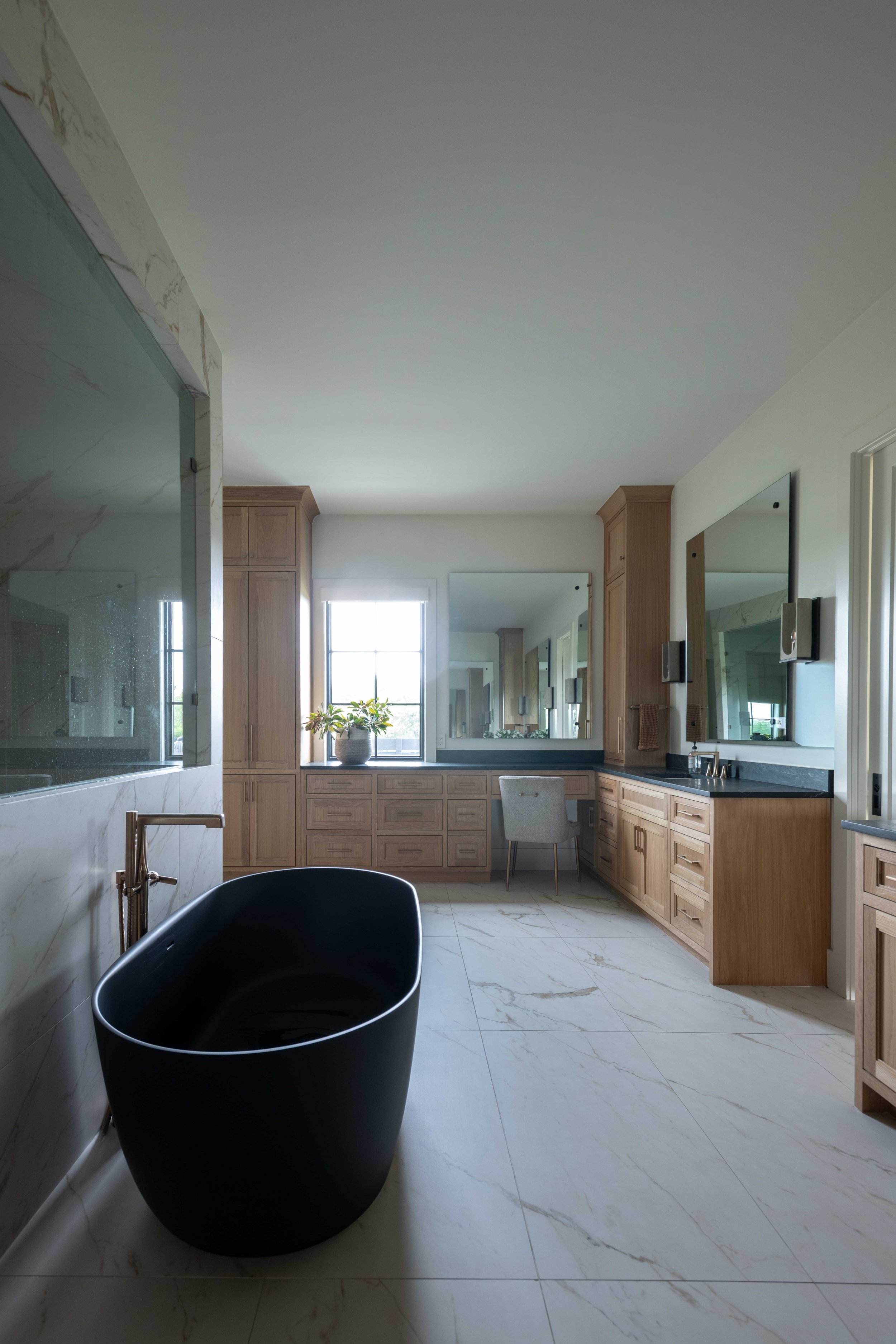 Modern bathroom with large mirrors, wooden cabinetry, and a black bathtub in the foreground. Natural light from a window illuminates the space. Marble tile flooring and walls add elegance.