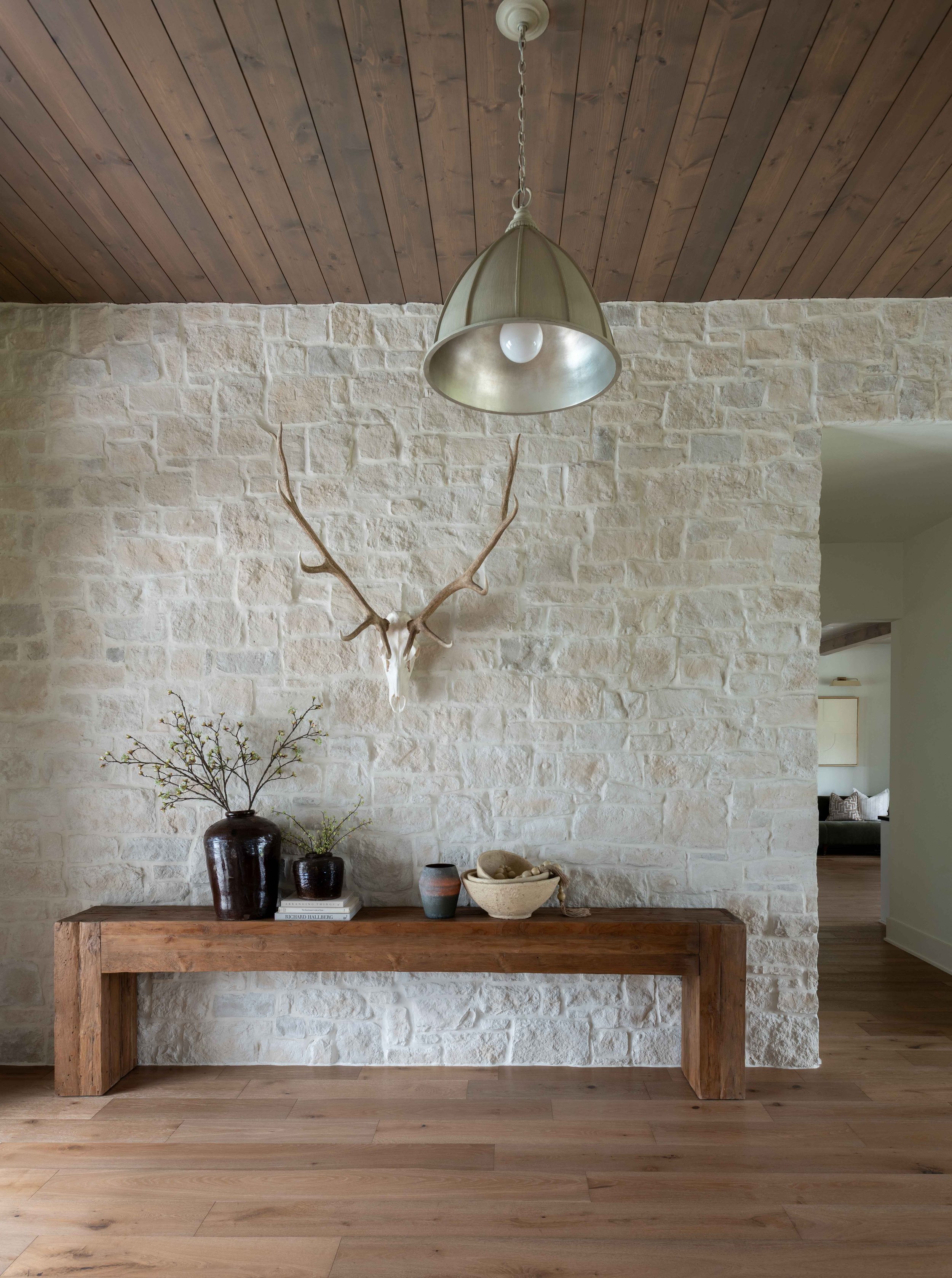 A decorative wooden table against a white brick wall with a mounted antler, three vases with plant arrangements, a bowl, and books on it. A wooden ceiling and a doorway leading to another room are visible.