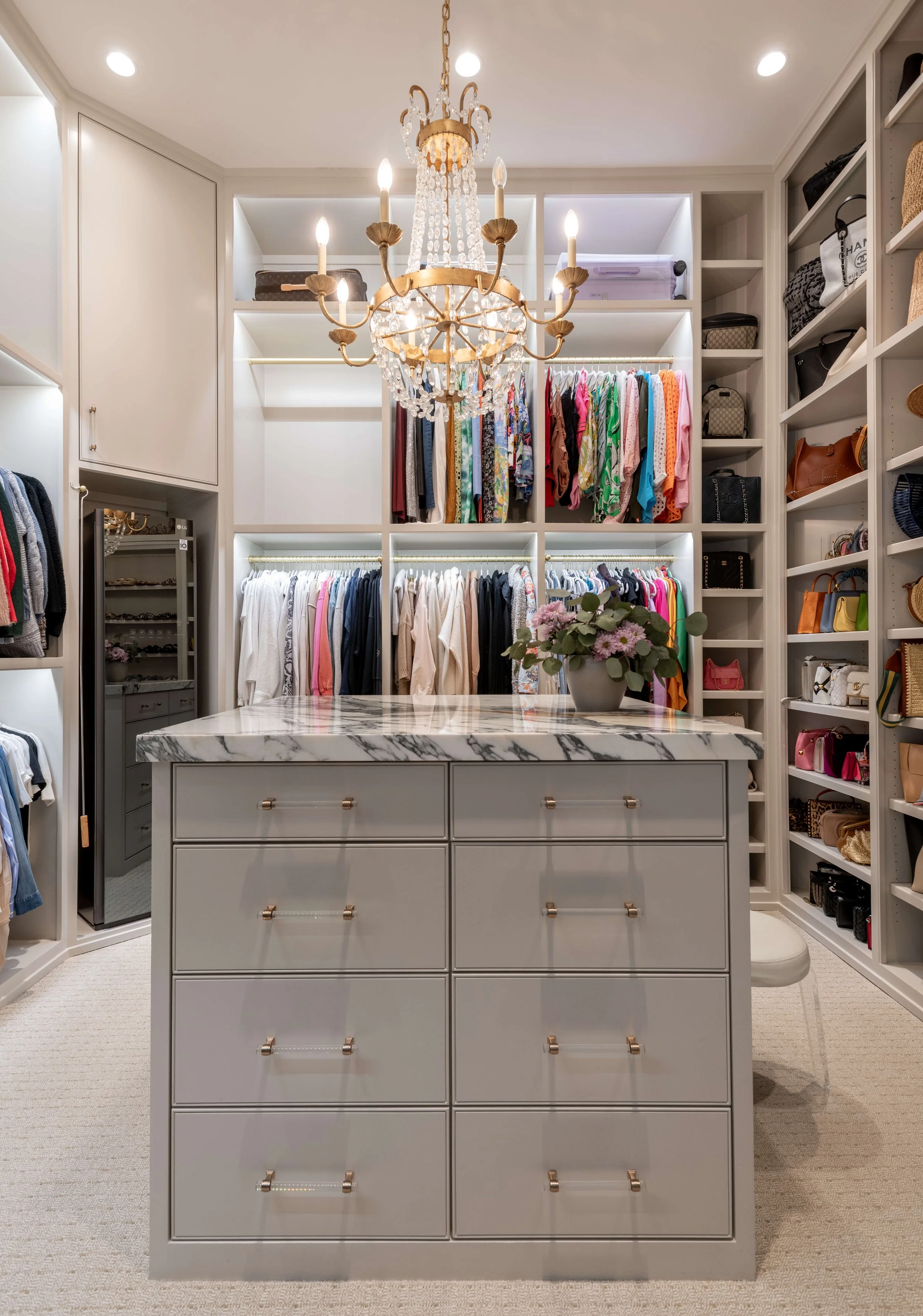 A walk-in closet with white shelving, organized clothes, handbags, and a marble-topped dresser. A chandelier hangs from the ceiling, and a vase with flowers is on the dresser.