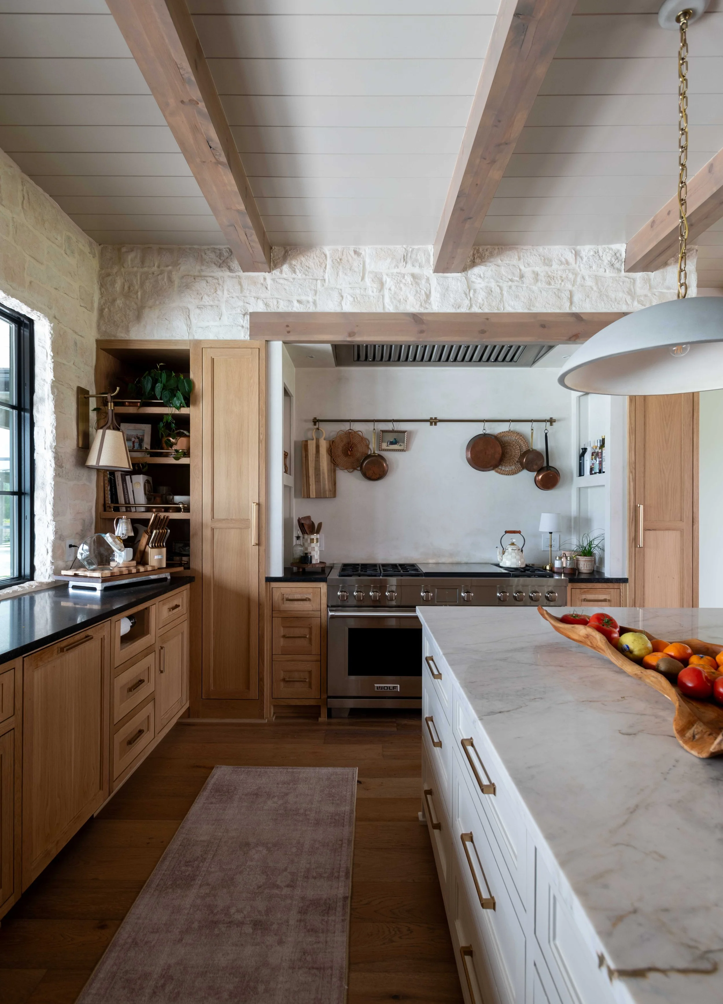A modern kitchen with wooden cabinets, a marble island with a wooden bowl of fruit, and a metal stove. The ceiling has white panels with exposed wooden beams, and a hanging light fixture is on the right. Copper pots hang on a rail on the wall.