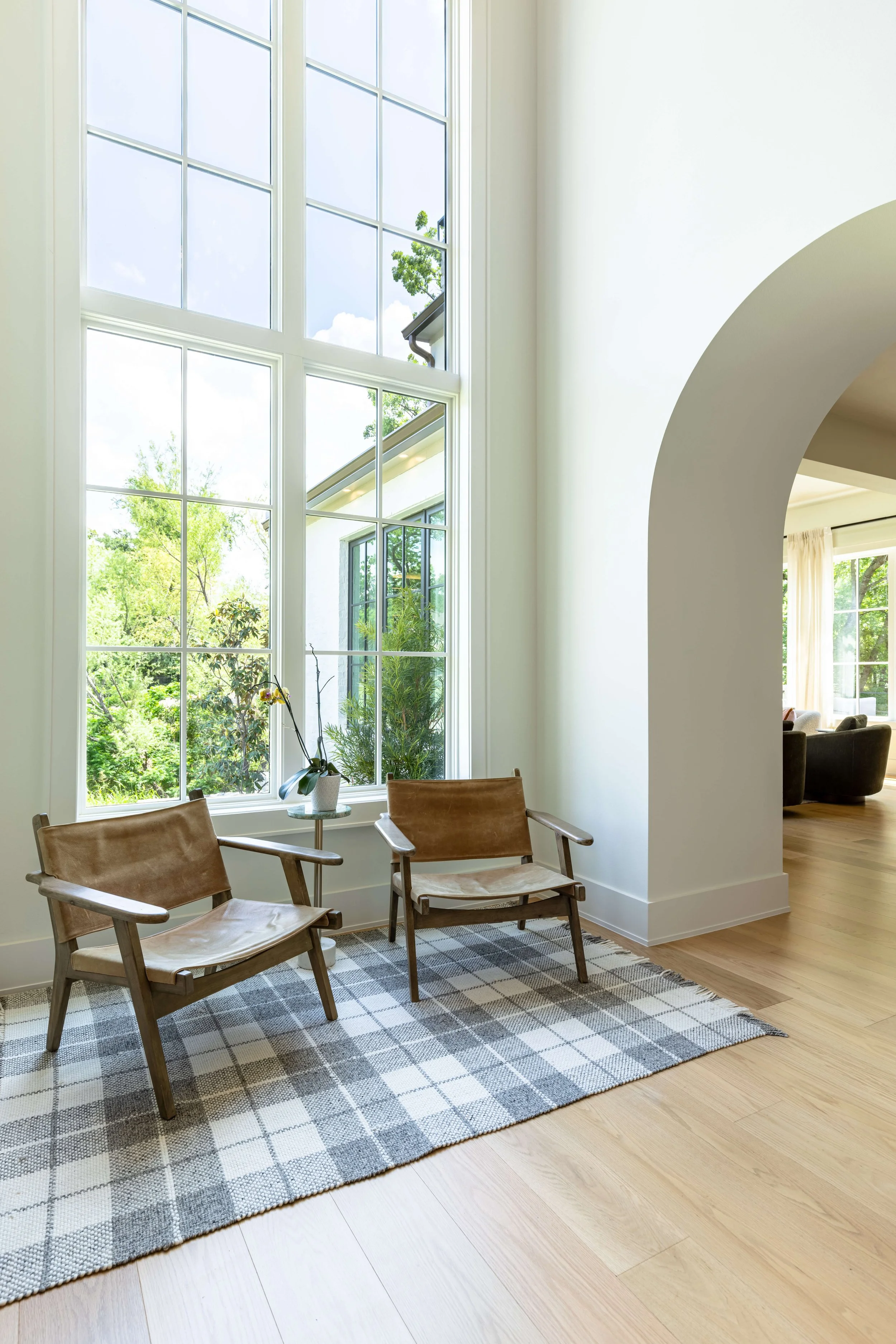 A bright living room with large windows showing a green outdoor landscape, two wooden chairs with leather seats and armrests, a small round table with a potted plant, a checkered rug, and a view into another room with a sofa and curtains.
