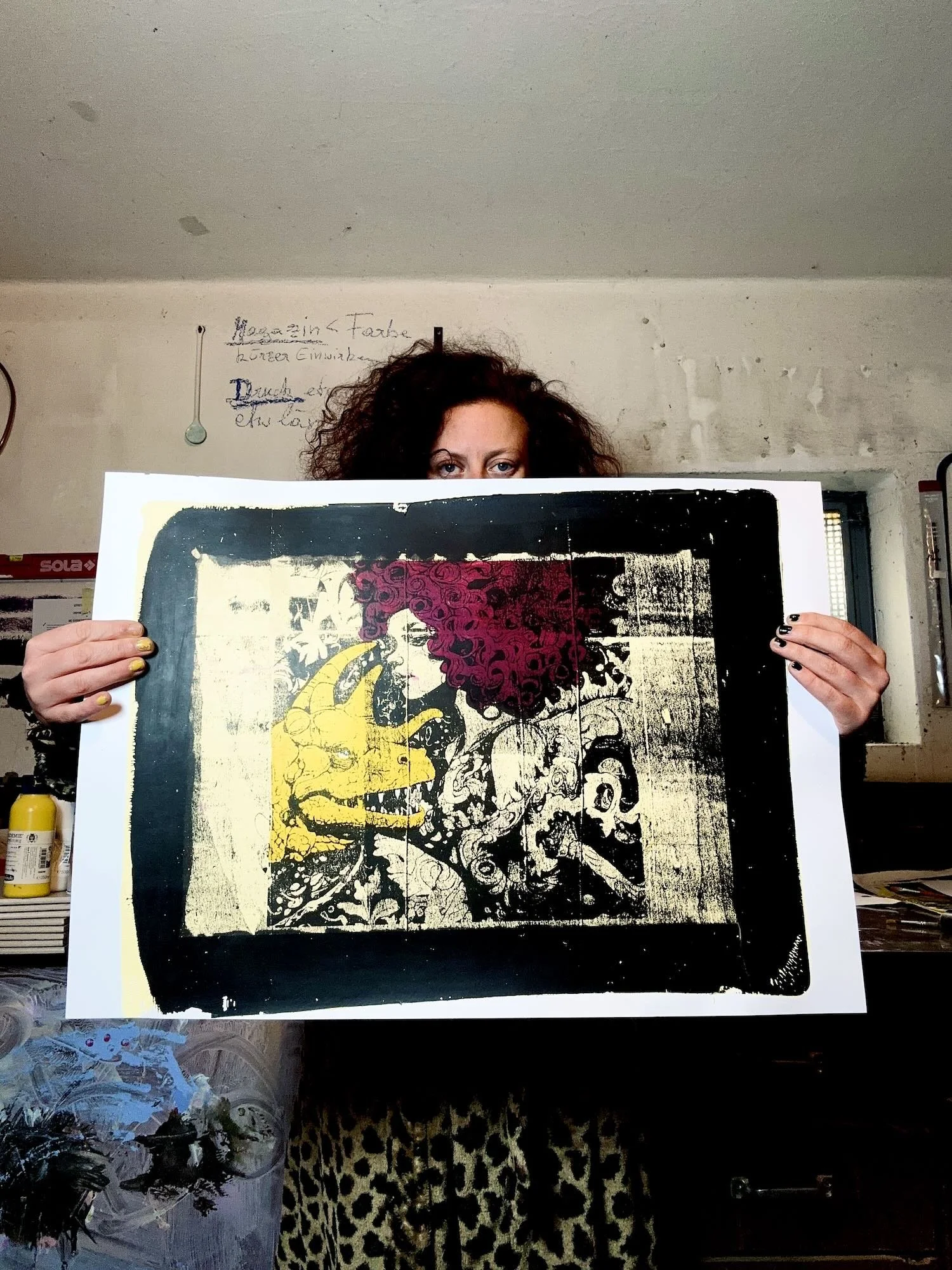 Eine Person mit lockigem braunem Haar hält ein großes Kunstwerk vor ihr, auf dem eine bunte, abstrakte Illustration mit gelber, pinker, schwarzer und weißer Farbe zu sehen ist. Die Person ist nur teilweise sichtbar, mit Fokus auf den Augen.