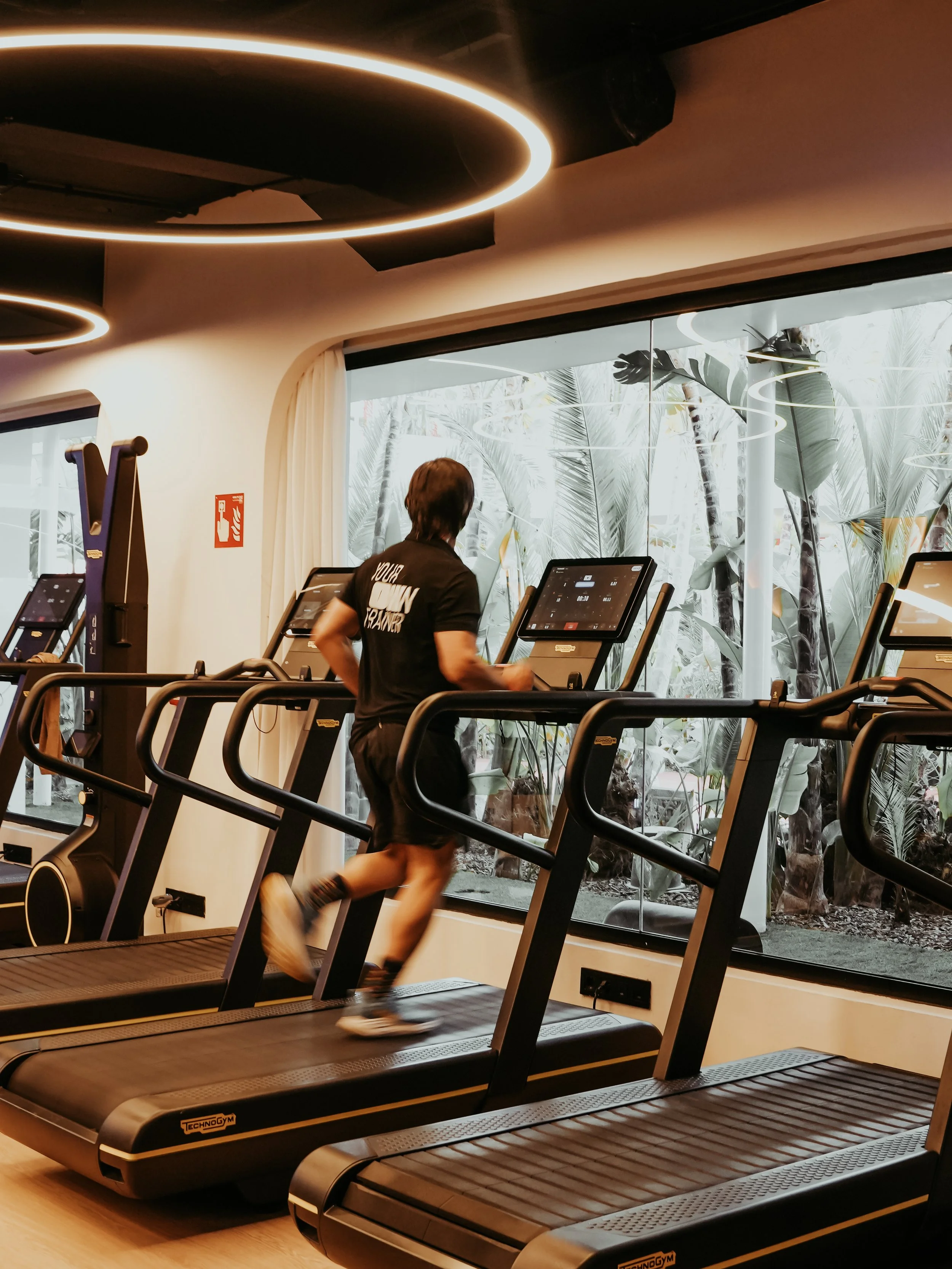 A person running on a treadmill in a gym, with large windows showing outdoor trees and plants in the Own Fitness Studio in Ibiza.