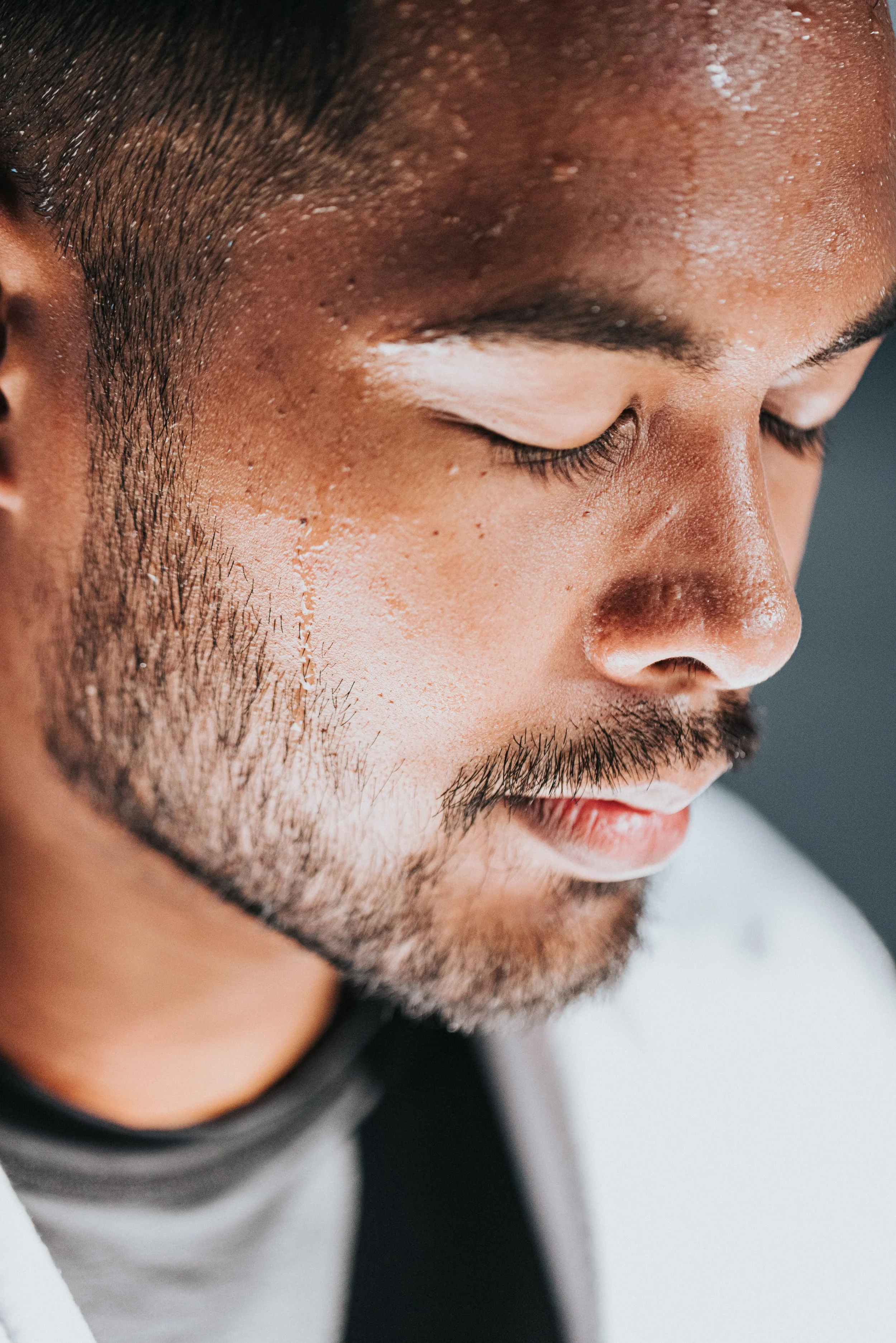 Close-up of a young man's face with closed eyes, showing facial hair, smooth skin, and short dark hair.