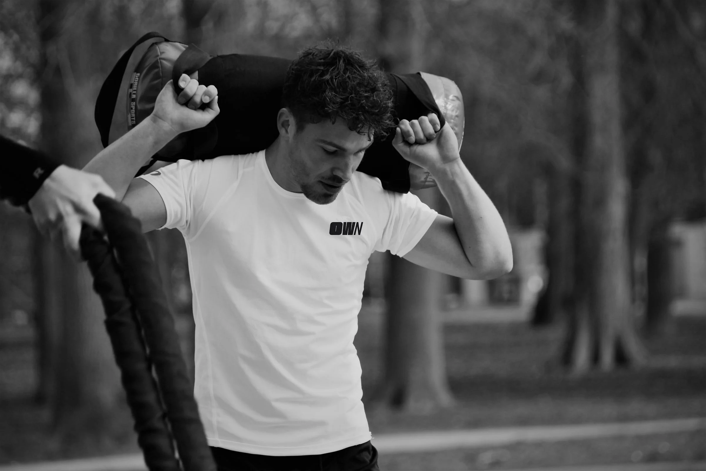 A young man with curly hair looks down, carrying a large duffel bag over his shoulders, outdoors with a blurred background.