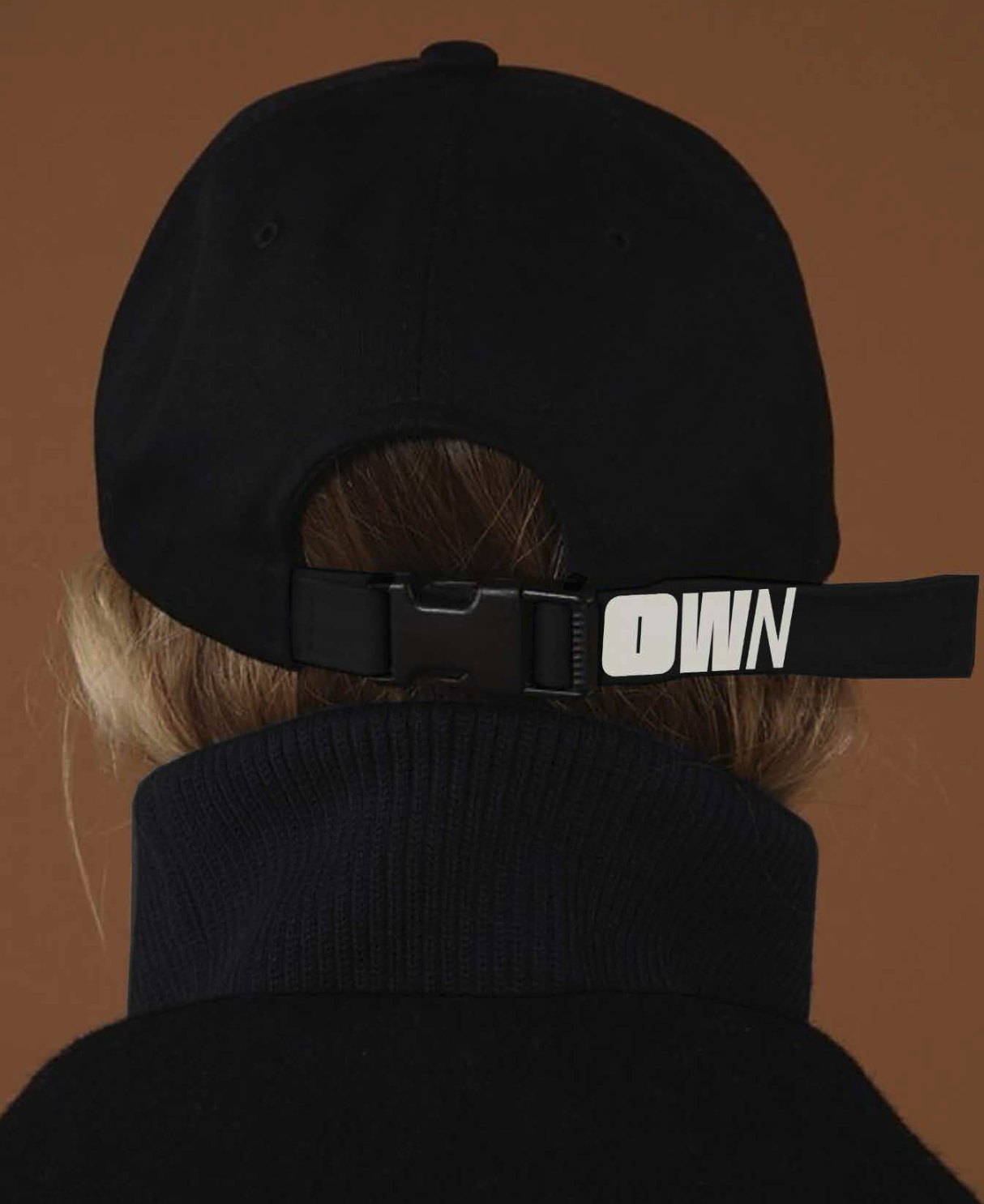 Back of a person wearing a black baseball cap with 'OWN' on the strap and a black turtleneck.