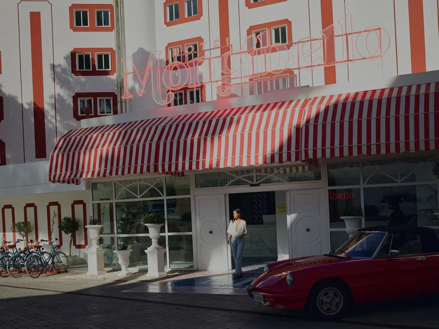 A woman stands outside a building with a red and white striped awning. There are potted plants and bicycles near the entrance, with a red sports car parked in front. The building has a neon sign that reads 'MOTELGIRL' and appears to be in an urban area.