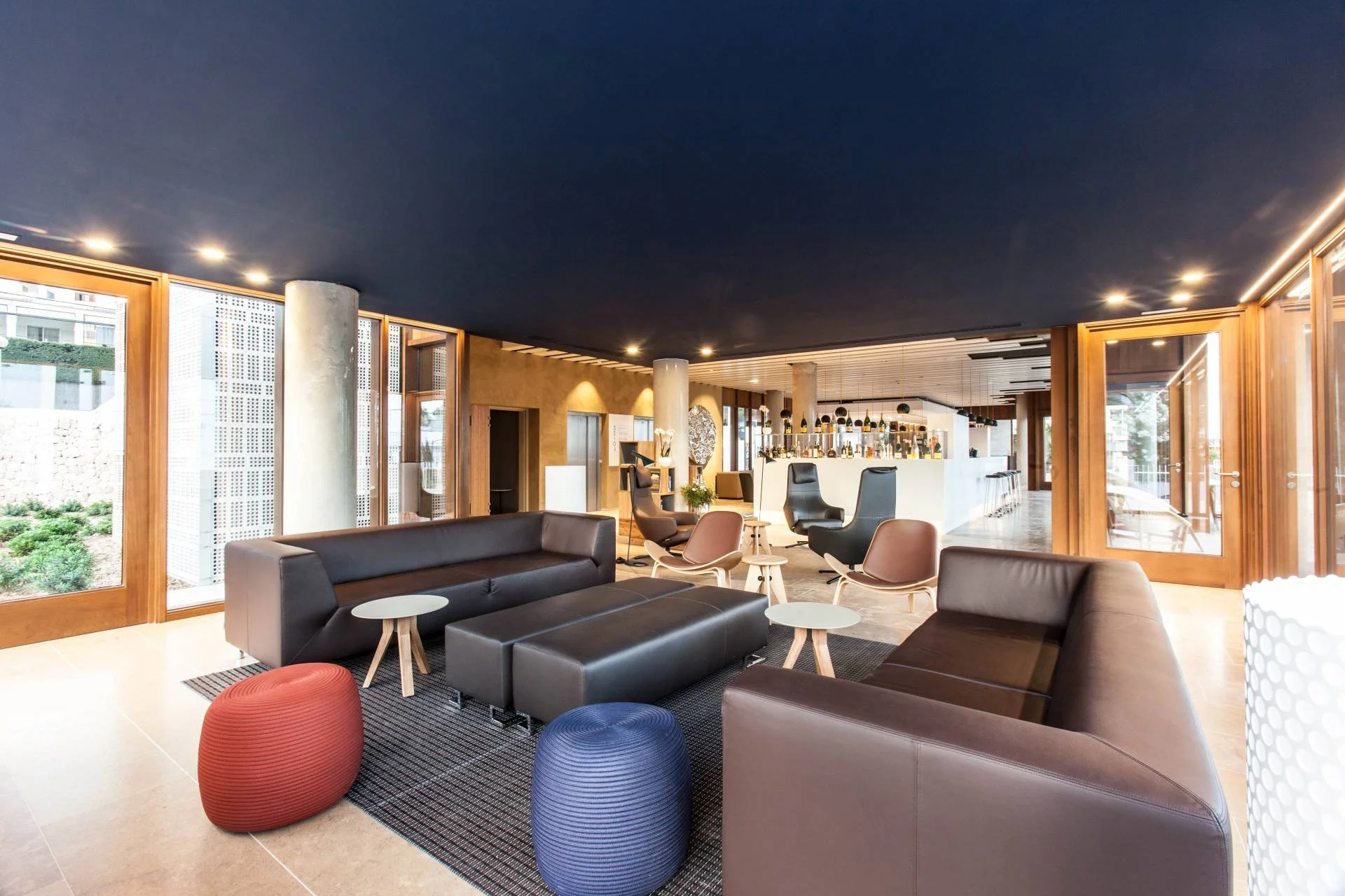 Modern hotel lobby with black leather sofas, colorful ottomans, small tables, and a bar area in the background with hanging pendant lights and bar stools.