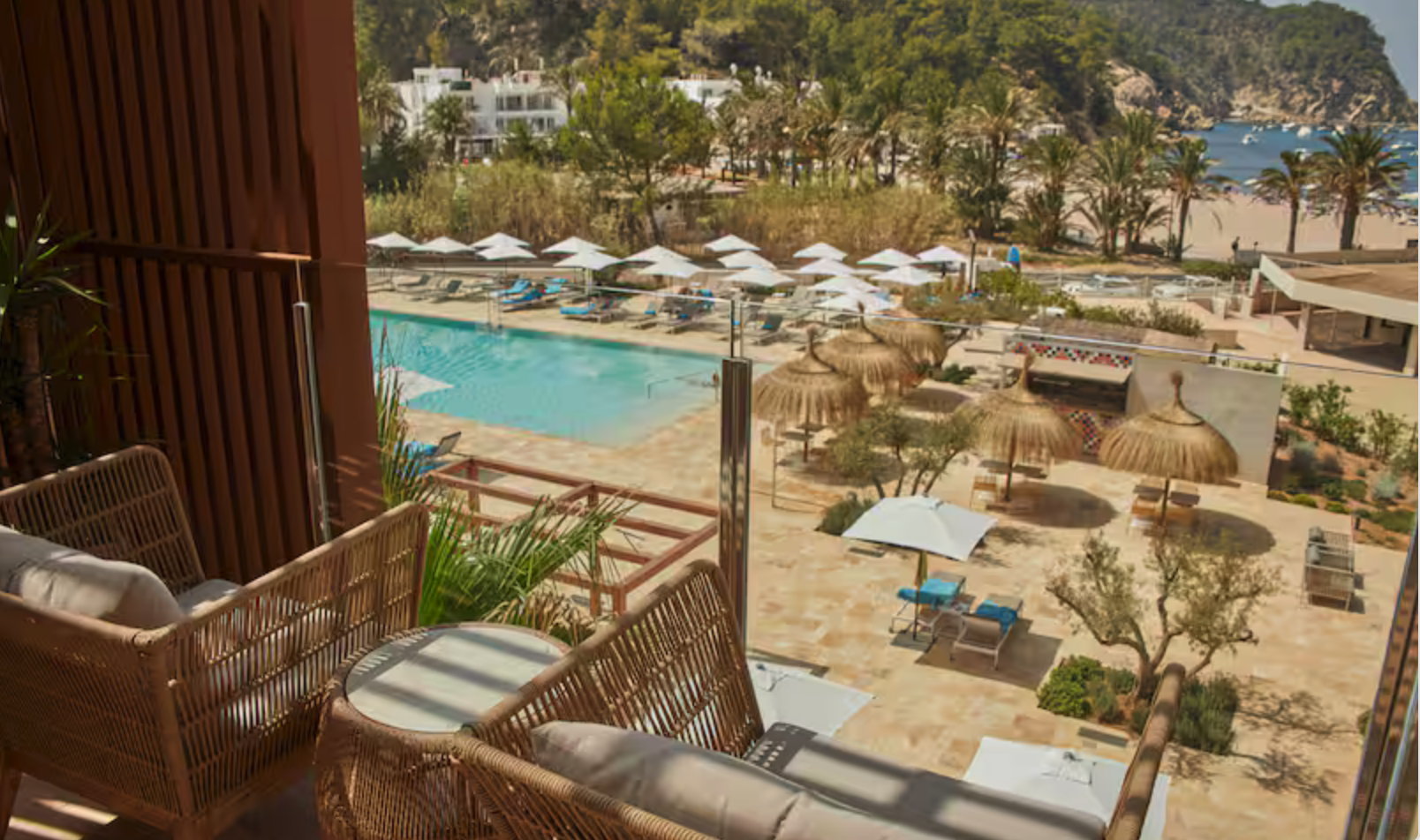 Balcony view overlooking a swimming pool area with sun loungers, umbrellas, and thatched-roof shaded seating, surrounded by palm trees with a sandy beach and ocean in the background.