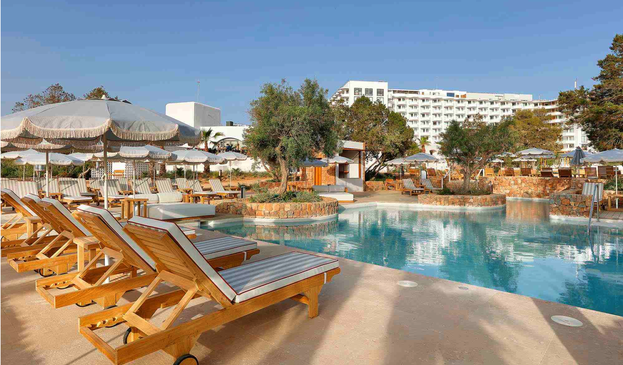 A luxurious outdoor hotel pool area with wooden lounge chairs featuring striped cushions, surrounded by white patio umbrellas. There are trees and a multi-story white building in the background under a clear blue sky.