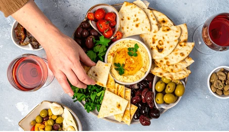 Verschiedene Zutaten für eine Meze-Platte, darunter Fladenbrot, Oliven, Tomaten, rotes Pfeffer, verschiedene Käsesorten, Hummus mit Petersilie, Wein und kleine Schälchen mit Kapern.